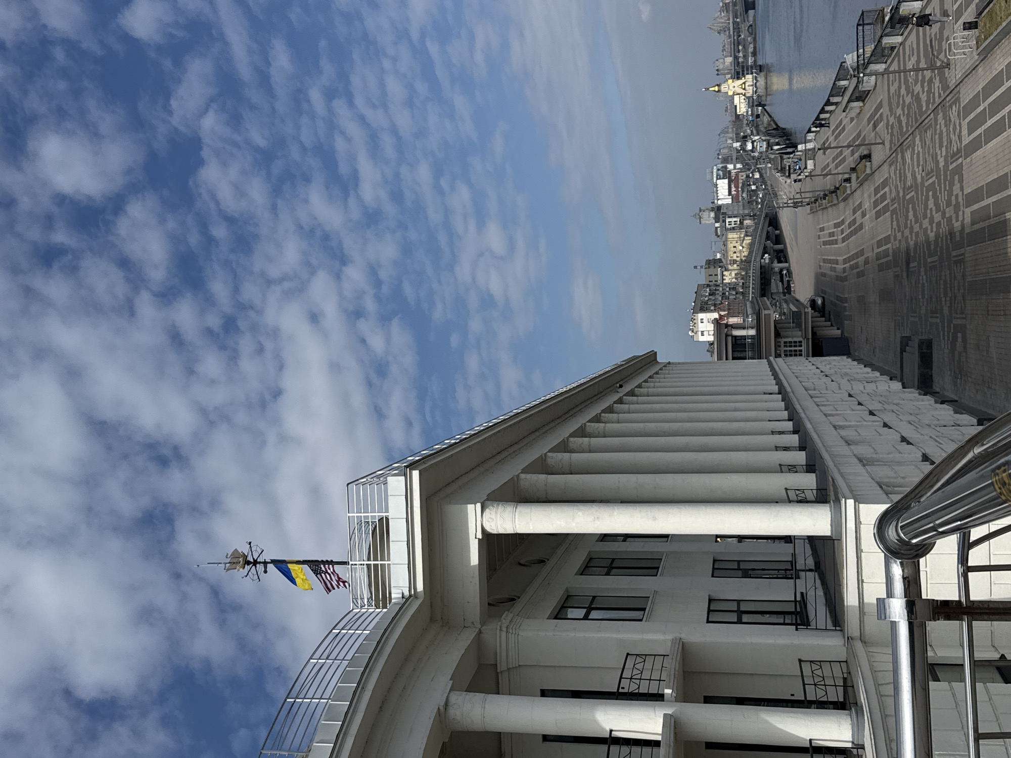 The image displays the Kyiv River Port (River Station) building and its adjacent embankment along the Dnieper River in Kyiv, Ukraine. The dominant structure is a grand, white, multi-story building featuring a prominent colonnade of numerous large, round columns supporting an upper level with a white railing. Dark-framed windows and black metal railings on lower balconies are visible on the building's facade. A chrome-finished metal railing occupies the lower-left foreground.
On the rooftop, a flagpole displays a Ukrainian flag (blue over yellow) positioned above an American flag (red, white, and blue), both partially unfurled. A decorative white ship model is mounted atop the flagpole. The sky above is blue with scattered white cumulus clouds.
To the right of the building, a wide, patterned paved promenade extends into the distance, running parallel to the Dnieper River. The promenade is equipped with lampposts, benches, and a black metal railing separating it from the water. Several individuals are present on the promenade, with one person visible walking towards the right in the mid-ground. The Dnieper River flows to the right, featuring several barges and boats docked along the embankment. In the distance, urban buildings, bridges, and the distinctive golden-domed St. Nicholas Church on the Water are visible across the river under a light atmospheric haze.