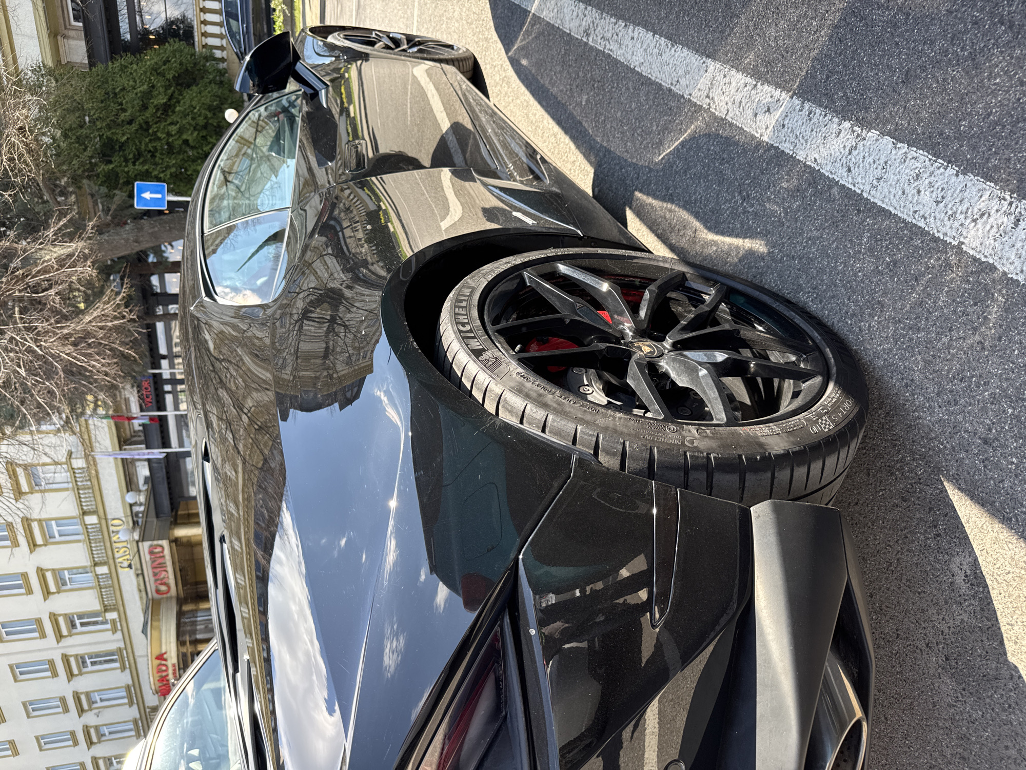 The image shows a black Lamborghini parked on a street in Plovdiv, Bulgaria. The vehicle is next to a white lane marking on the asphalt. The reflection on the car's surface shows buildings, including one with a visible "Casino" sign. A blue directional sign is visible in the background. The car's rear wheel is prominently displayed, showing the tire and the Lamborghini logo on the wheel hub.