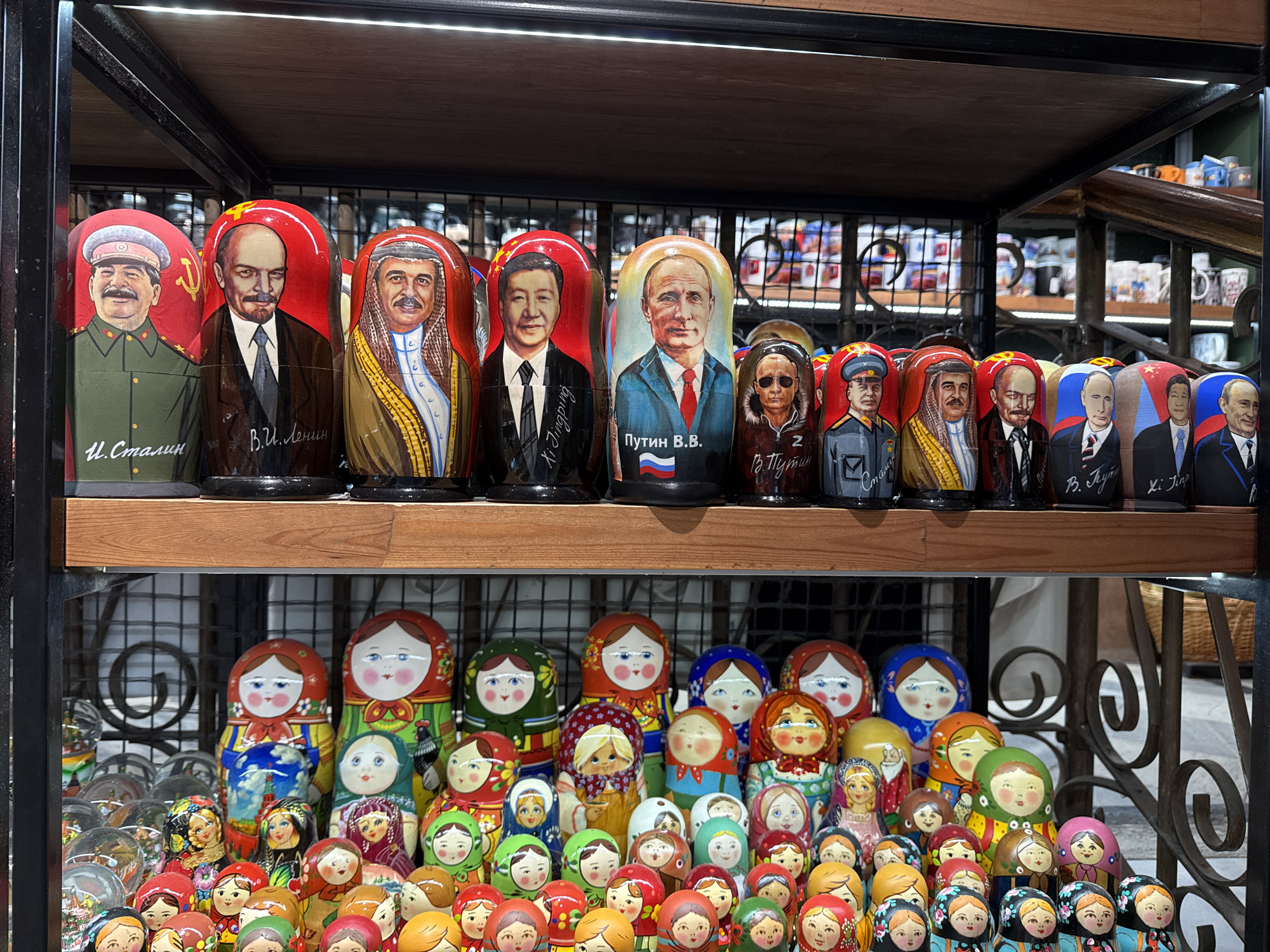 In an indoor market stall, a shelf displays a collection of Matryoshka dolls. The dolls are arranged in two rows. The top row features larger dolls depicting famous political figures. From left to right, the dolls are of Joseph Stalin, Vladimir Lenin, a figure identified as "Xi Jinping" (likely Xi Jinping), Vladimir Putin, and two other figures. The dolls are painted with the figures' faces and distinctive attire. The bottom shelf is filled with smaller, traditional Matryoshka dolls, which are painted in various colorful patterns and depict women in traditional Russian dress. Some of these dolls have red headscarves, while others have different colored headscarves and clothing. The overall environment suggests a souvenir shop or a market selling traditional Russian crafts. The lighting is artificial, casting a slight glare on the dolls. The background consists of shelves with other merchandise, including mugs and decorative items. The location is identified as Unknown, Unknown.