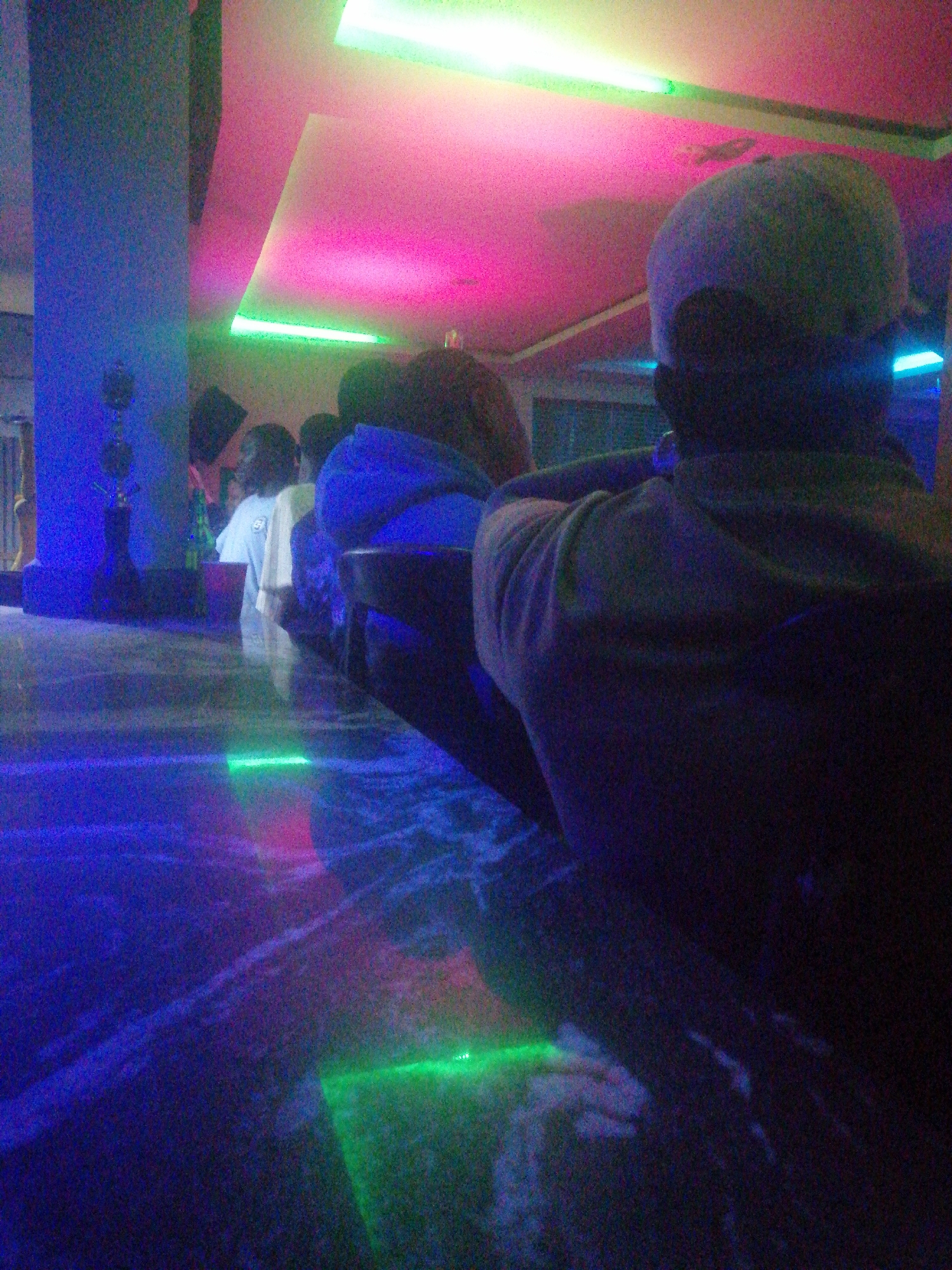 The image depicts an indoor scene, likely a bar, club, or lounge, characterized by vibrant and colorful artificial lighting during the evening or night. The perspective is from behind a person seated at a long counter, looking into the venue.
In the immediate foreground, a person is seen from the back, wearing a light-colored baseball cap and a dark shirt or hoodie, with their arms resting on the counter. Stretching across the lower half of the frame, the reflective counter surface is illuminated by various colored lights, primarily deep blue and green, with subtle pink reflections. Several bright green light strips are visible on or embedded in the counter.
Further along the counter, several other individuals are partially visible, mostly from behind or the side, gathered as if socializing. One person in the mid-ground appears to be wearing a light-colored t-shirt. On the far left, a tall pillar is prominently lit with a strong blue glow.
The ceiling and upper wall areas are adorned with bright pink and green strip lights, which cast a luminous, neon-like ambiance across the entire space. The overall dimness of the environment, punctuated by these intense light sources, signifies a nighttime setting. The image exhibits noticeable digital noise or grain, common in low-light photography. There is no visible text in the scene. The setting strongly suggests a social gathering or entertainment event.