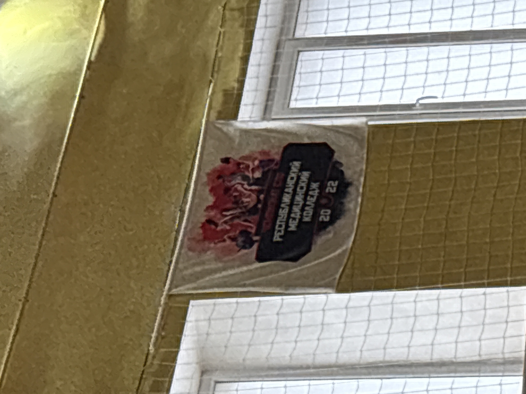 The image shows a sign hanging in front of a window in a building in Izhevsk, Russia. The sign contains the words "Республиканский Медицинский Колледж 2022". The window has a metal grate on it. The area outside the window is not visible.