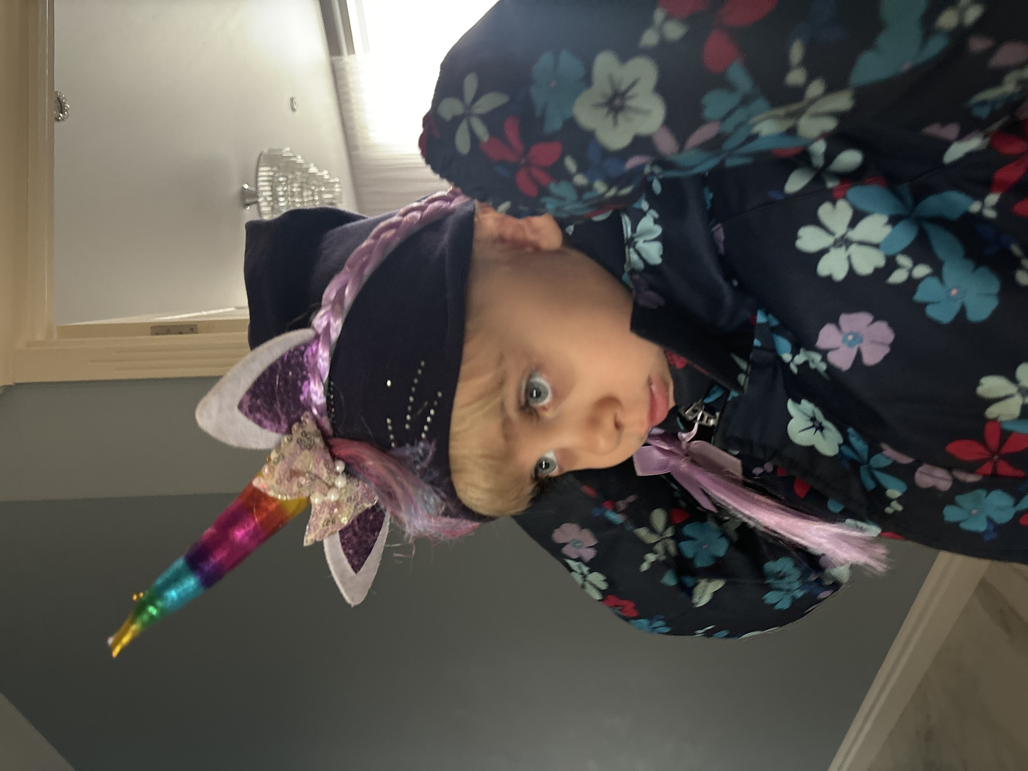 A young child with fair skin and blue eyes is wearing a dark blue hooded coat with a floral pattern. The child is also wearing a unicorn headband with a multicolored horn and sparkly ears. The background is a blurry interior scene with white walls and a light fixture. The city and location are unknown.