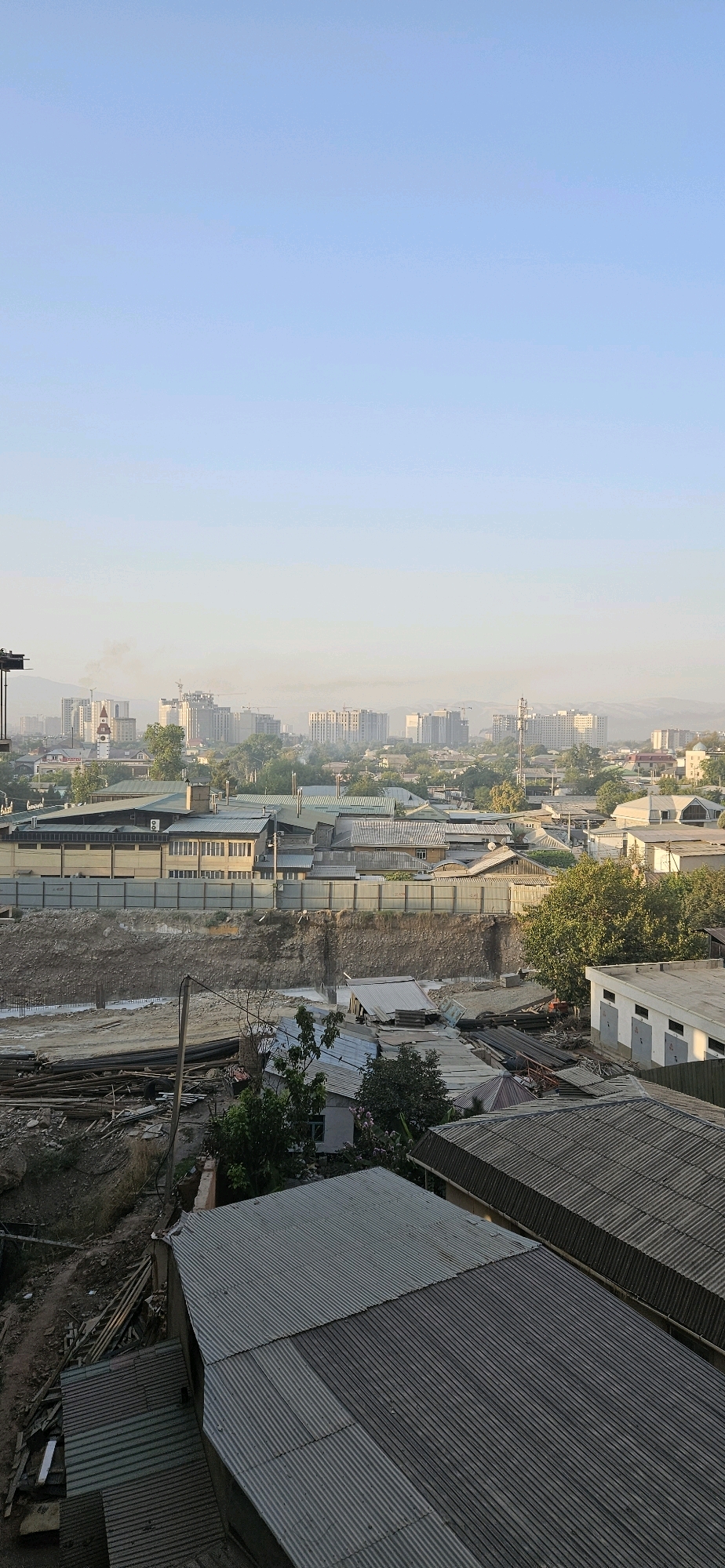This is a high-angle, vertical, outdoor shot of a city under a hazy sky, likely taken in the morning or evening due to the soft light and lack of distinct shadows. The foreground is dominated by the corrugated metal roofs of several low-rise buildings, appearing to be industrial or storage structures. These roofs have varying patterns and colors, some appearing weathered.
In the midground, a large construction site is visible. It features a deep excavation with visible concrete foundation work, surrounded by a gray metal barricade. Piles of lumber, pipes, and other construction debris are scattered around the site. Behind the construction area, a row of more buildings with metal roofs and some with light-colored facades can be seen, separated by a line of trees.
The background reveals a sprawling cityscape with numerous multi-story buildings, some appearing to be apartment blocks or office buildings, extending towards the horizon. The buildings are varied in height and architectural style, with some under construction indicated by cranes. A thin layer of haze or smog partially obscures the distant city and mountains. The sky is a pale, clear blue, transitioning to a lighter hue near the horizon. The overall impression is one of urban development with ongoing construction and a densely built environment.
Based on visual cues like architectural styles of the buildings and the general urban landscape, the image is consistent with an area within the city of Dushanbe, Tajikistan. Specifically, the combination of low-rise industrial buildings, construction sites, and distant apartment blocks is characteristic of some developing districts within the city.