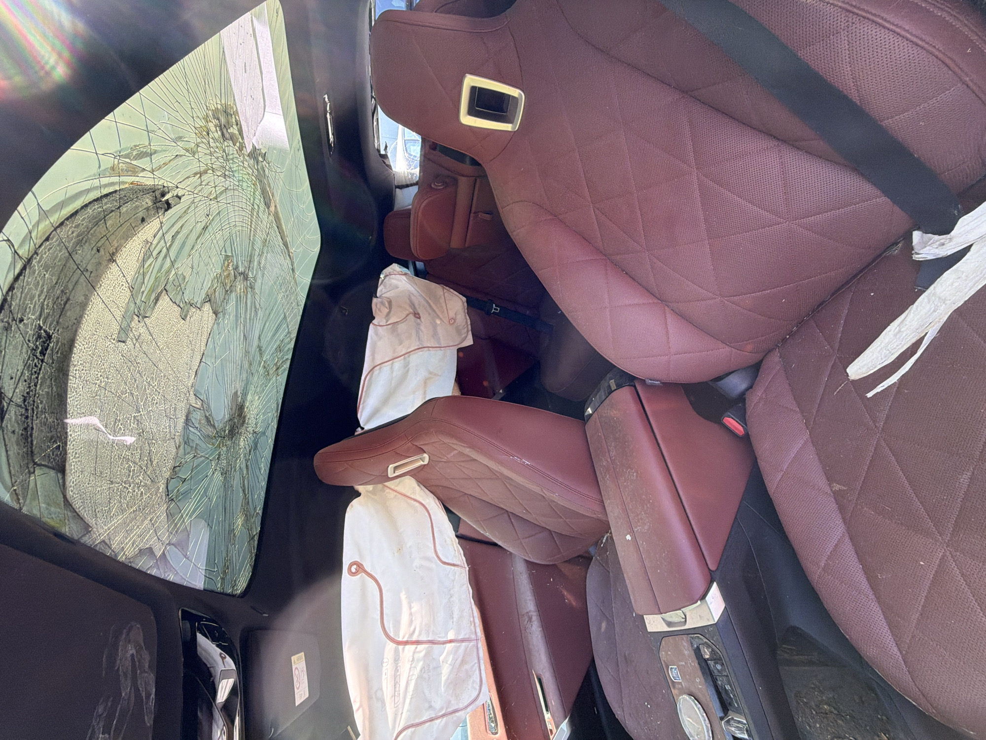 The image shows the interior of a damaged car, likely in a junkyard or salvage area, in Burgas, Bulgaria. The panoramic sunroof is heavily shattered, with glass fragments spread across the interior. The airbags for the front seats have deployed, indicating a significant impact. The car's interior features red leather seats, which appear dusty and possibly soiled. Debris is scattered on the seats and the center console. The overall impression is one of a vehicle that has sustained severe damage.