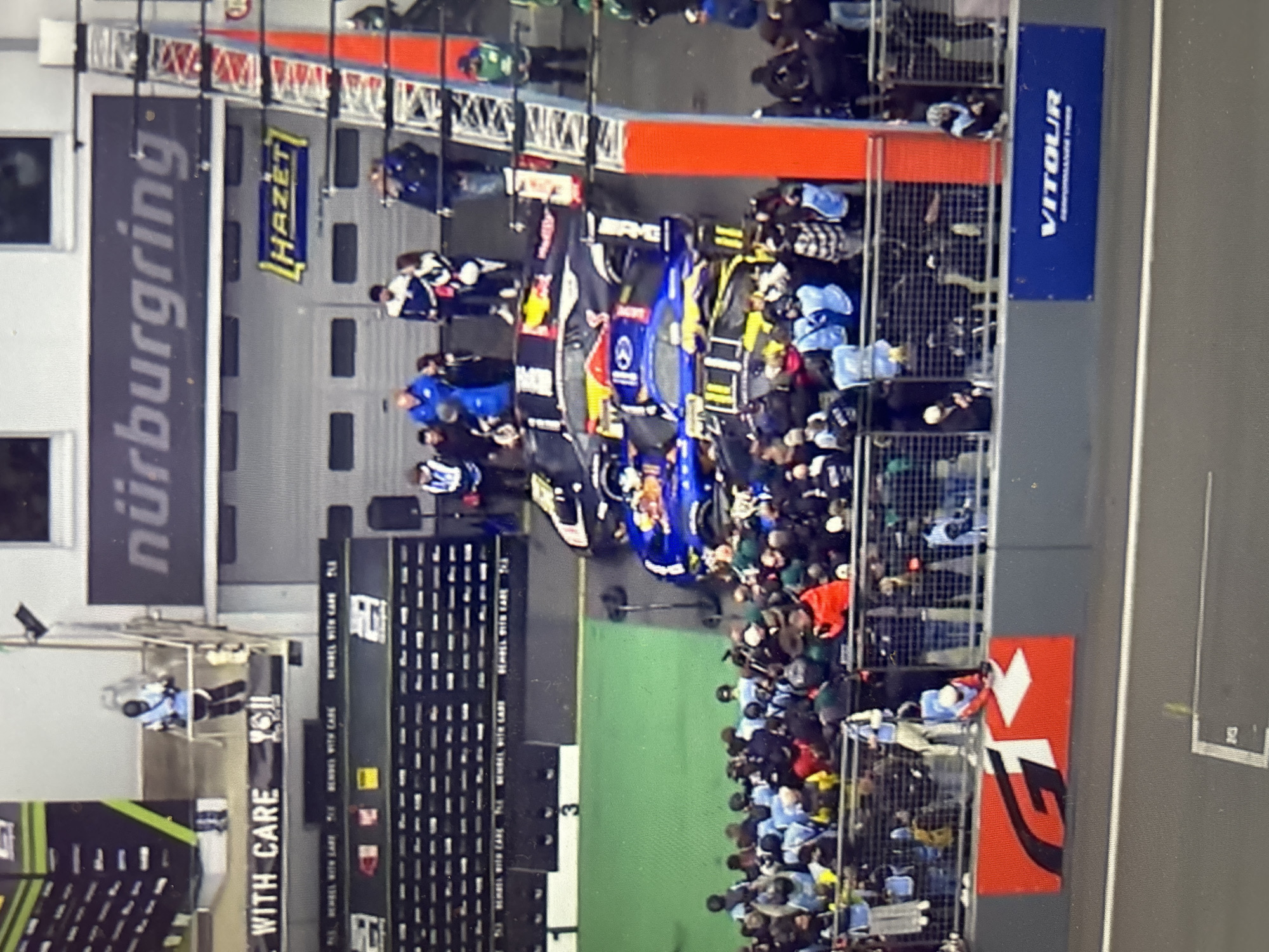 The image depicts an overhead view of a race event, likely a motorsport competition, taking place at the Nürburgring, evidenced by the prominent "nürburgring" signage. Several race cars, including one with Red Bull livery, are lined up on a track. A large crowd of spectators is visible behind fencing and barriers. Advertising banners with brand names such as "HAZET", "VITOUL" and "REMMEL WITH CARE" are also visible. The overall environment suggests a professional racing event. The image does not contain any information suggesting that the location is in the city of Sofia, Bulgaria.