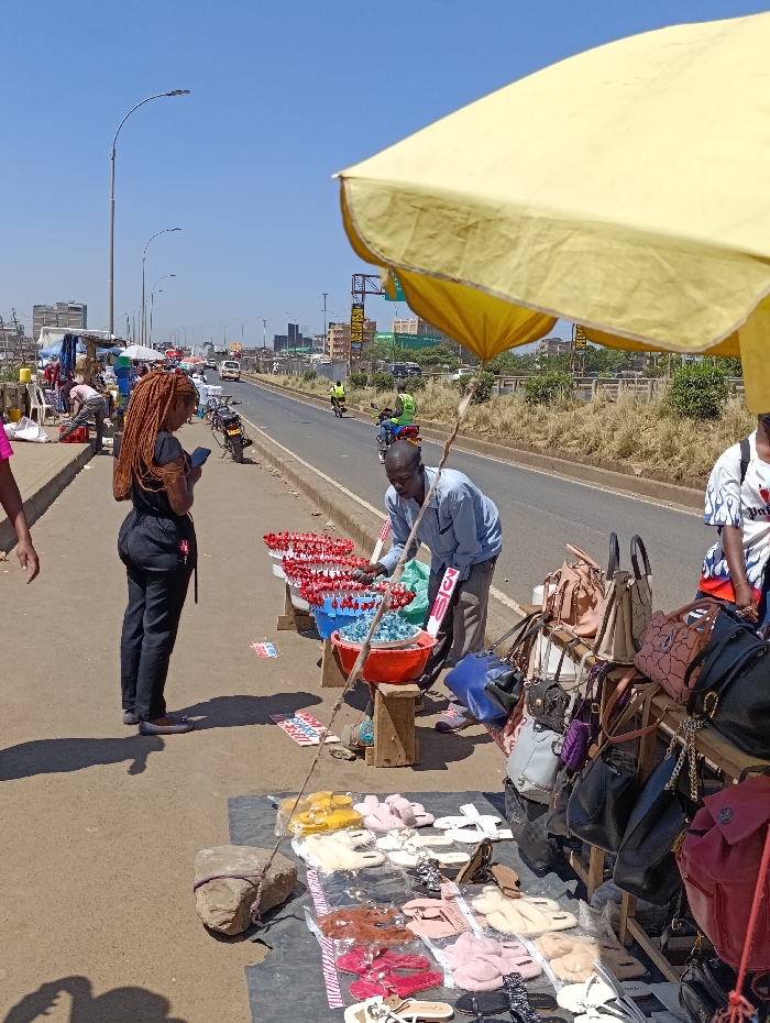 The scene is a street market in Githurai, Kenya, located at the coordinates -1.2 latitude and 36.921 longitude. A man is selling various items including sandals and handbags under a yellow umbrella. He is interacting with a woman who is looking at the sandals. There are other vendors selling goods and several pedestrians walking by. The environment is an urban street with a paved road and sidewalks. There are buildings and trees in the background. The sky is clear and blue. The sun is shining brightly. The scene suggests a typical day of commerce and social interaction in Githurai.