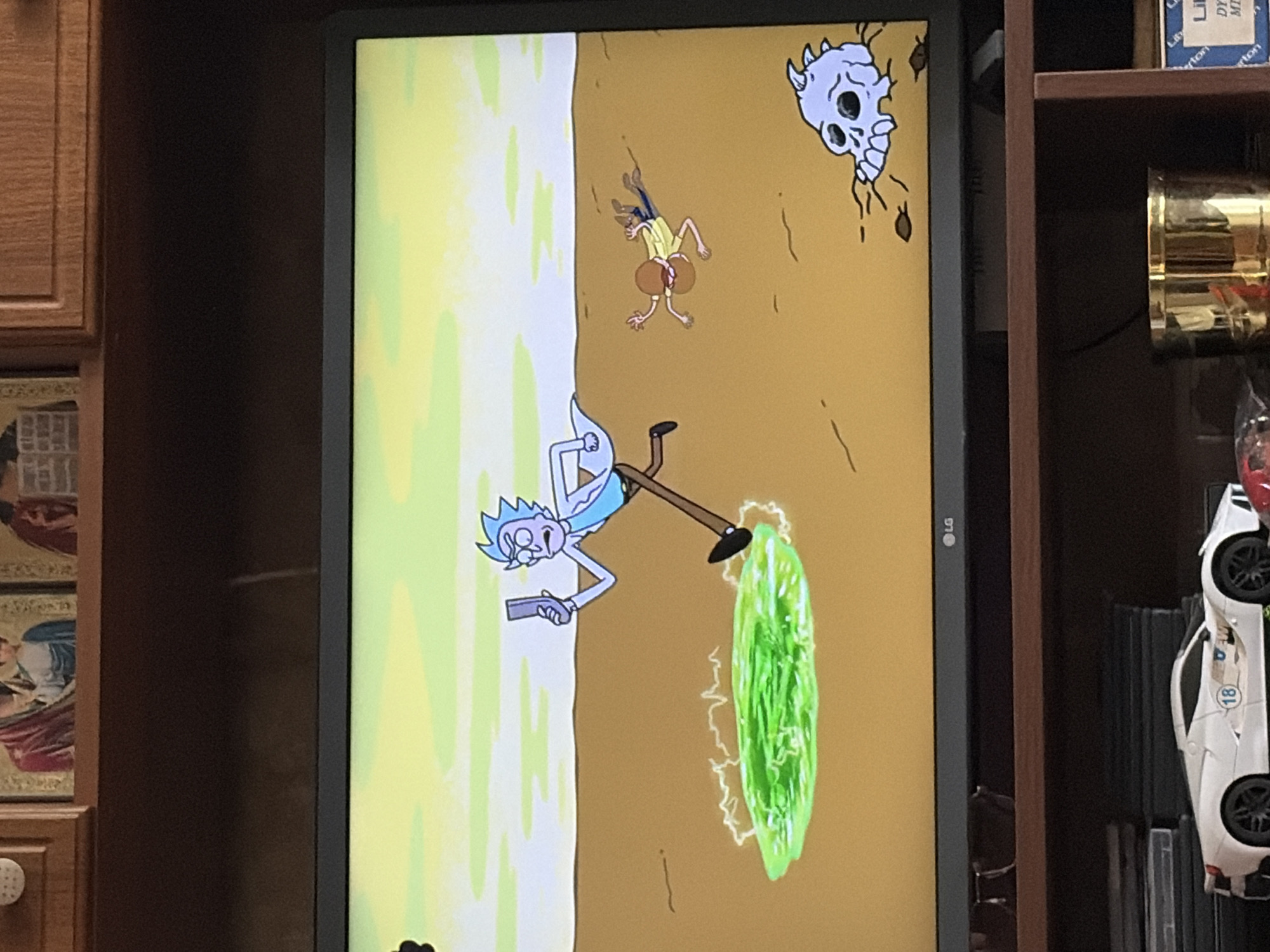 The image shows a television screen displaying a scene from the animated show Rick and Morty. In the foreground of the screen, Rick Sanchez is running towards a green portal. To the right of Rick, another character, possibly Morty Smith, is lying on the ground. In the bottom right corner of the screen, there is a cartoonish skull. The television is placed on a shelf in what appears to be a living room or entertainment area. On the shelf below the television, there are several objects, including a white toy car with the number 18 on it, a brass container, and some other miscellaneous items. The background above the television shows a wooden cabinet with religious icons.