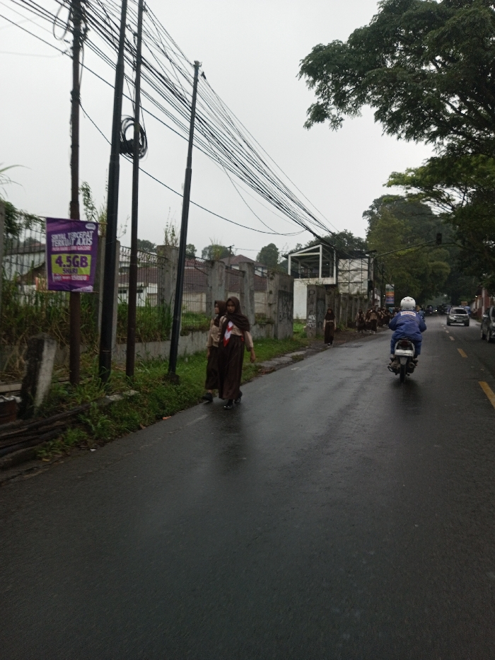 The scene is captured in the city of YonPhoogla, Bhutan, on an overcast day with wet asphalt indicating recent precipitation. The image depicts a two-lane road with a yellow dividing line, extending into the distance where multiple vehicles are present. On the left side of the road, three utility poles are positioned, heavily strung with numerous black electrical and communication cables. A purple banner advertising "4.5GB" and "AXIS" is affixed to one of these poles. Adjacent to the road is a narrow strip of grass, followed by a concrete fence, parts of which are covered in graffiti. Beyond the fence, residential structures and dense foliage are visible.
Two individuals, appearing to be young females, walk together on the left edge of the wet road, dressed in matching brown skirts, light-colored tops, and brown headscarves/hijabs. Further down the road, additional individuals in similar attire are also visible near the fence line. A person wearing a white helmet and a blue jacket rides a white scooter away from the viewer on the wet road. Tall green trees line the right side of the road, and an unfinished or renovated building structure with exposed white framing is visible on the left side, further down past the initial fence section. The sky is uniformly grey and overcast.