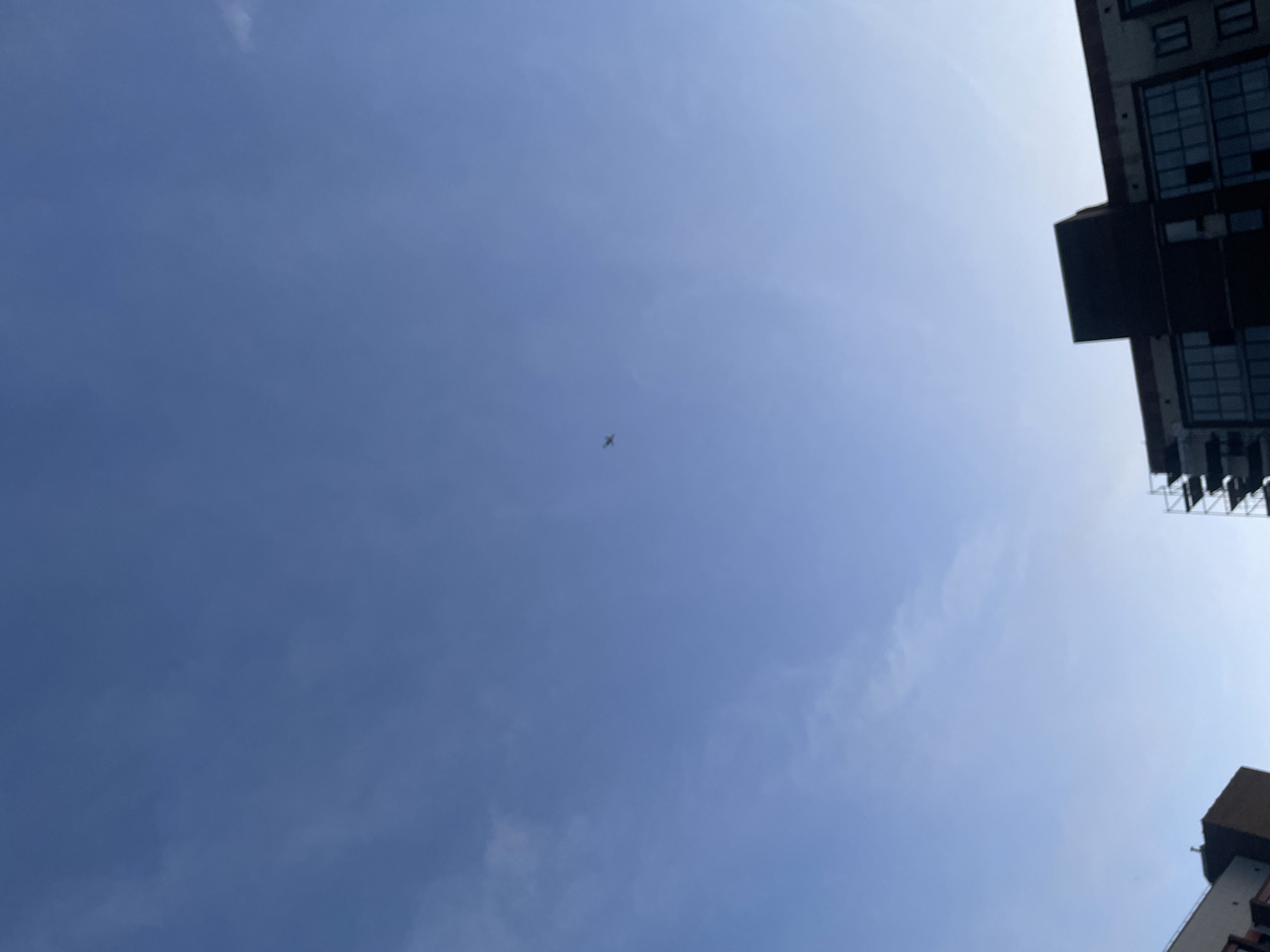 This media file shows a view looking up at a clear blue sky. A small airplane is visible in the distance, flying through the sky. On the right side of the image, the top of a modern building with many windows can be seen. The building's architecture suggests an urban environment. The bright sky indicates it is daytime, likely midday, under fair weather conditions. The composition creates a sense of looking up from ground level between buildings. There is no visible text in the image.