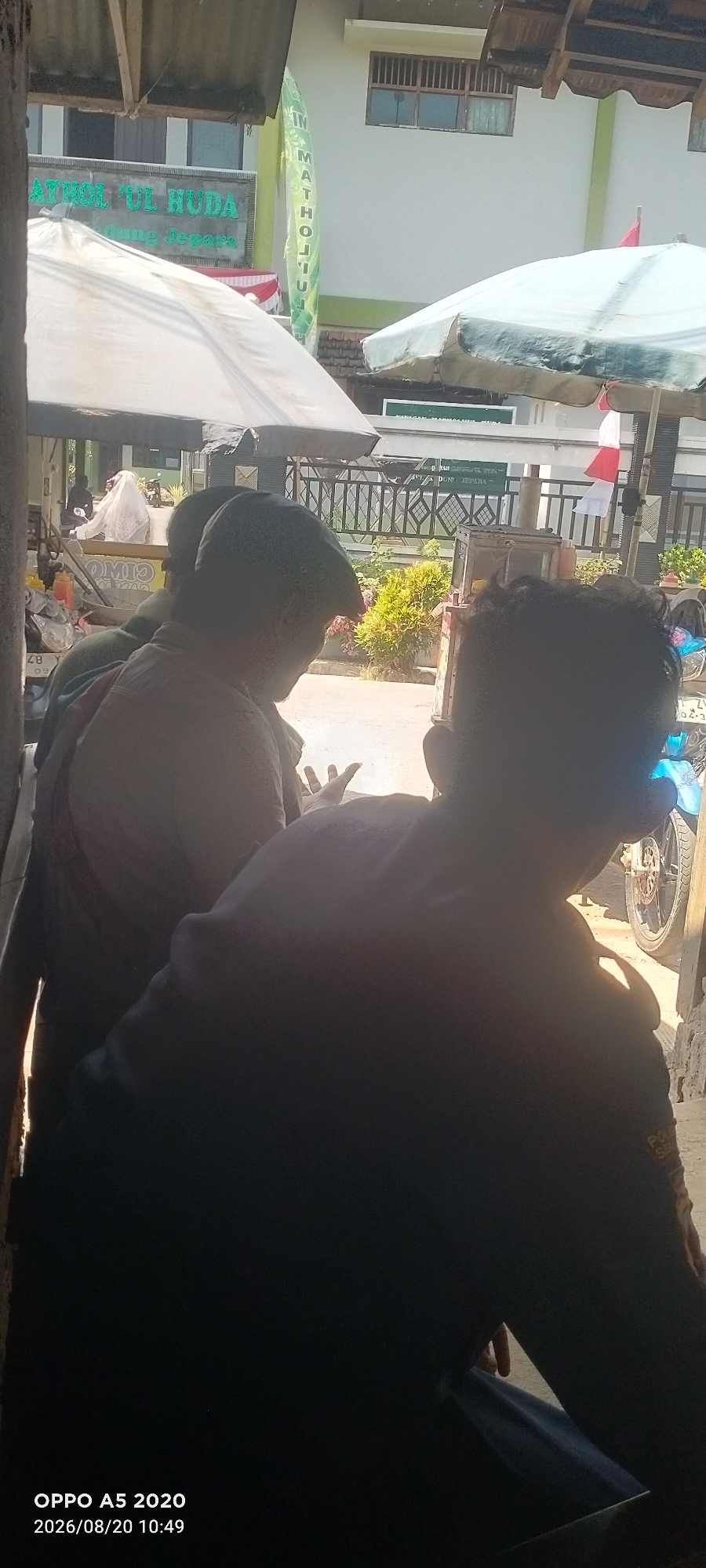 The image shows a street scene in Bugel, Indonesia. In the foreground, three individuals are partially visible, with their backs to the camera. The person on the right is wearing a dark shirt with a patch on the sleeve that reads "POL." The person in the middle is wearing a light-colored shirt and a cap. The person on the left is wearing a patterned shirt. They appear to be engaged in conversation or observing something in front of them.
In the background, there is a building with the words "MATHOL'UL HUDA" and "MADRASAH" visible on a banner. Two white umbrellas are set up, possibly for a stall or shelter. A motorcycle is parked on the right side of the image. The overall environment suggests a typical street scene in a semi-urban area. The timestamp at the bottom of the image indicates the photo was taken on August 20, 2020, at 10:49 AM.