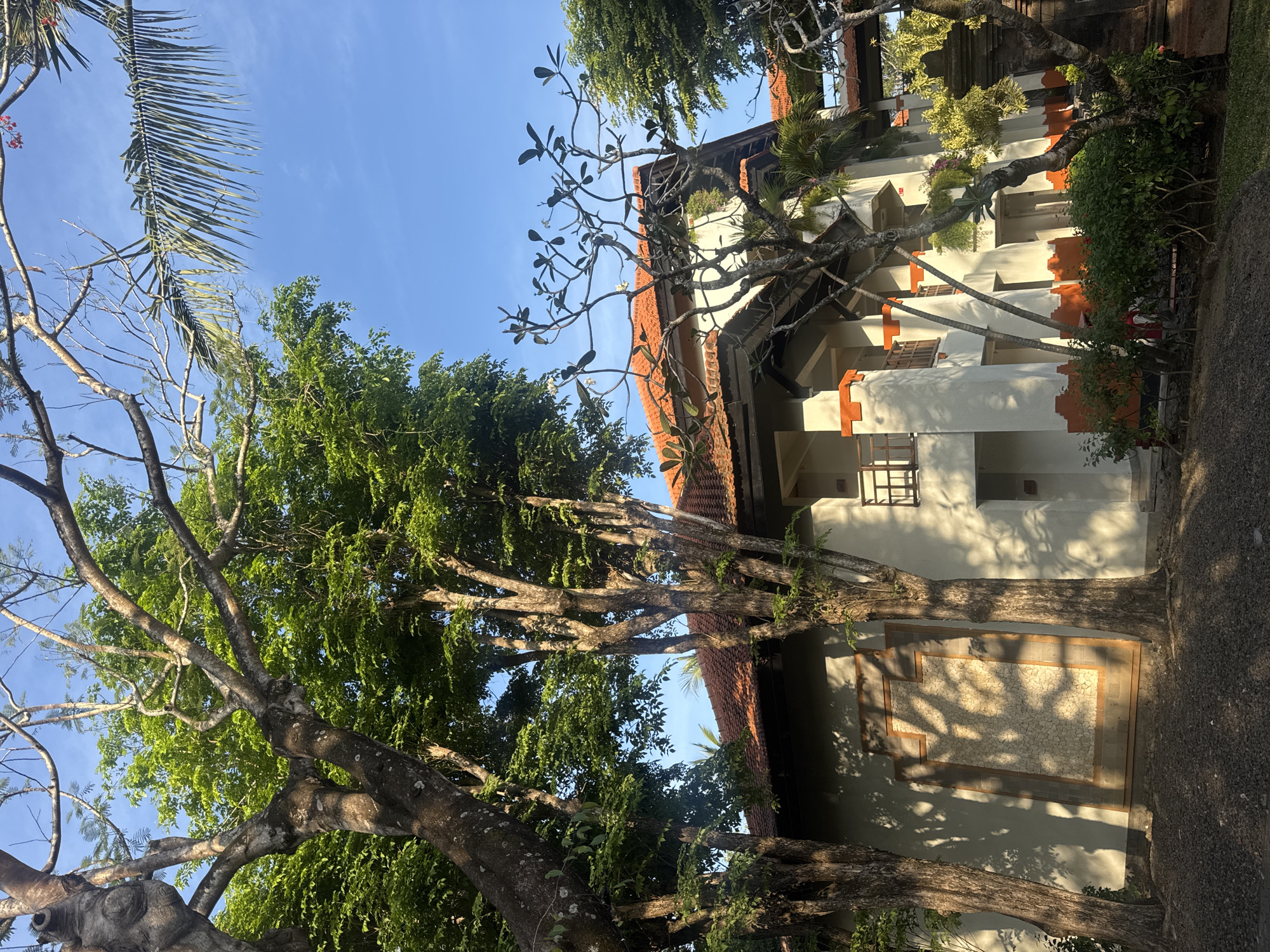The image depicts the exterior of a building with traditional Balinese architecture in Kuta Selatan, Indonesia. The building has white walls, terracotta tile roofs, and orange accents. It is surrounded by lush green trees and foliage, with a clear blue sky in the background. The sunlight casts shadows of the trees onto the building's walls, creating a dappled effect. In the foreground, there is a gravel path and some bushes. The scene appears peaceful and serene, likely a resort or hotel complex.