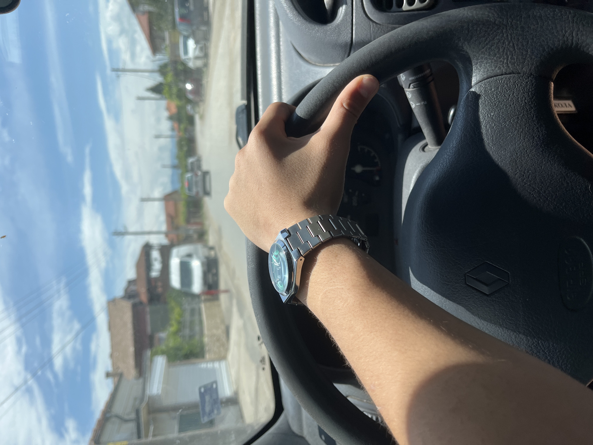 The image, rotated 90 degrees clockwise, captures a first-person perspective from inside a vehicle, looking out through the windshield. In the immediate foreground, a person's right hand grips the upper left side of a black, textured steering wheel. A metallic wristwatch with a round, possibly green or blue, dial and a multi-link bracelet is prominently worn on the person's left wrist.
The steering wheel features a dark, geometric Renault logo on its center airbag cover. Behind the steering wheel, a control stalk for turn signals or wipers is visible, with some faint, indecipherable text on it. A section of the dashboard is also visible, and the word "KOLA" can be seen printed on a surface near the top right of the steering wheel.
Through the windshield, a bright, sunny outdoor scene unfolds. The sky is largely clear with scattered white clouds. The setting appears to be a residential street or road in Ivailo, Bulgaria, lined with various buildings, including houses with visible rooftops and facades. Several parked cars and possibly a utility truck are seen along the street. Utility poles and power lines crisscross the view, along with some trees and foliage. The strong sunlight suggests it is daytime, likely midday or afternoon. The person appears to be driving or about to drive the vehicle.