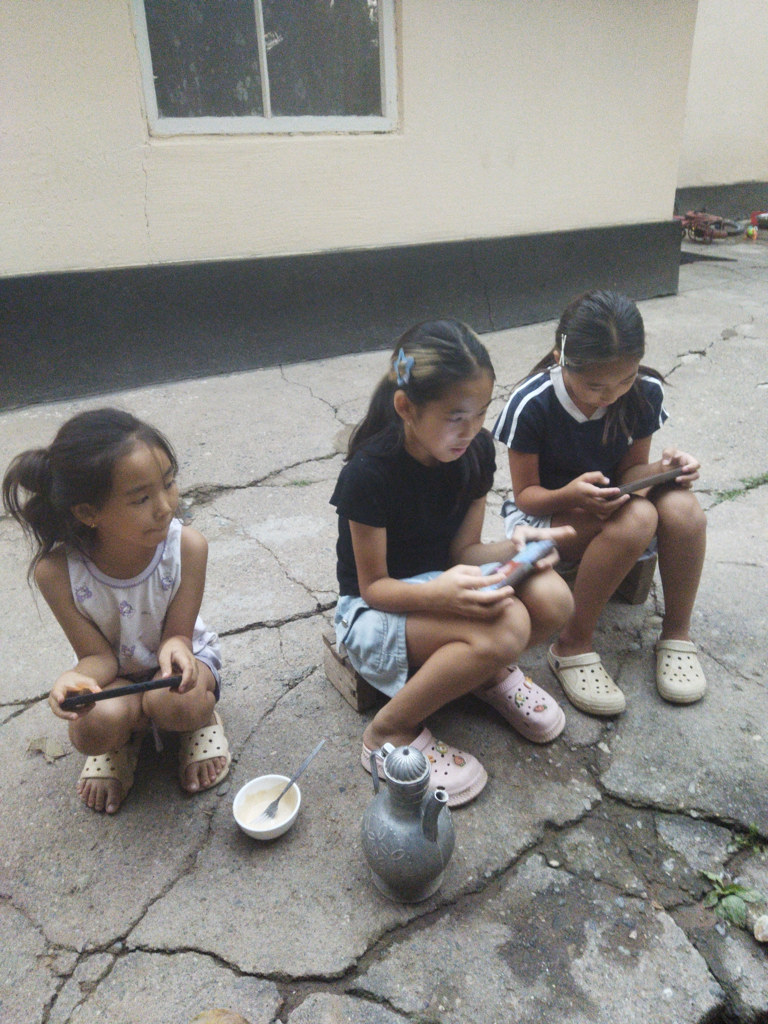 Three young girls are crouched on a cracked concrete ground in front of a building with a light-colored wall and a dark gray foundation. The girl on the left, wearing a white sleeveless dress with purple floral patterns and beige clogs, holds a dark object, possibly a phone, in her hands. Next to her is a small white bowl containing a pale yellow substance and a fork. To her right, a second girl, dressed in a black t-shirt with white stripes and denim shorts, is looking down at a mobile device in her hands. She is wearing beige clogs. The third girl, on the far right, is wearing a dark blue and white striped shirt, light-colored shorts, and beige clogs. She is also engrossed in a mobile device. In the background, a bicycle and other objects are visible. The overall scene suggests a casual outdoor setting in Mailuu-Suu City, Kyrgyzstan, where the children are engaged with their devices.