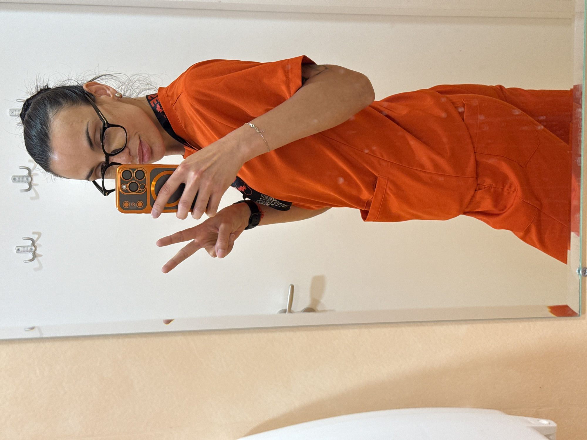 An indoor mirror selfie captured in Pleven, Bulgaria, shows a person with dark hair pulled back in a bun, wearing dark-framed glasses and a bright orange uniform, possibly scrubs. The individual is holding a smartphone with an orange case, taking the photo in the mirror. With their right hand, they make a peace sign, while the left hand holds the phone. A slight smile is visible on their face, suggesting a relaxed or casual mood.
The person wears a small silver stud earring, a thin silver chain bracelet on their left wrist, and a black digital watch on their right wrist. Around their neck, a dark lanyard is visible, bearing the partially legible text "GES". The uniform appears to be a two-piece set, with details like pockets visible on the trousers.
The setting appears to be a room with light-colored walls, possibly white or off-white. On the wall to the left, a set of white wall hooks is visible. The lower portion of the image shows a surface, possibly a countertop or a lower wall section, in a light beige or cream color. The mirror itself has some small white specks or dust marks on its surface. The overall lighting suggests an indoor environment without specific time-of-day cues.