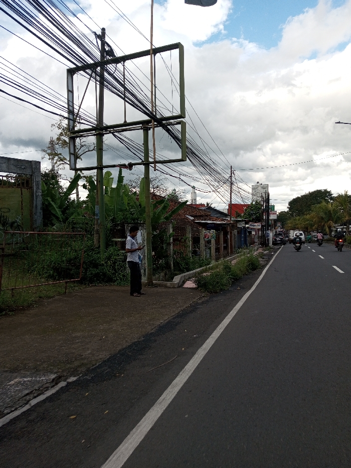 This scene is located in the city of Serpang Shalli, Bhutan. The image depicts a street view under a partially cloudy sky.
On the left side of the frame, a man stands on a dirt sidewalk, facing slightly right, looking down at a mobile phone held in both hands. He is wearing a light-colored, short-sleeved collared shirt, dark trousers, and sandals. Immediately behind him, and extending further left, is a dense accumulation of green vegetation, including large-leafed plants and overgrown bushes. A prominent, empty billboard frame, constructed from green metal beams forming two rectangular sections, stands on the left, partially obscuring the background. Numerous black electrical or communication cables are strung overhead between utility poles, many converging on the billboard structure, creating a dense network against the sky. Behind the vegetation and billboard, various single-story buildings with tiled roofs are visible, one featuring a white, minaret-like tower with a conical top in the distance. A rusted metal gate is partially visible on the far left.
The central part of the image is dominated by an asphalt road with a solid white line marking its left edge. Multiple vehicles, including several motorcycles with riders and passengers, and a white van, are traveling away from the viewer. The right side of the road is lined with additional buildings, commercial signage, and mature trees extending into the background. A street light pole is visible on the far right.