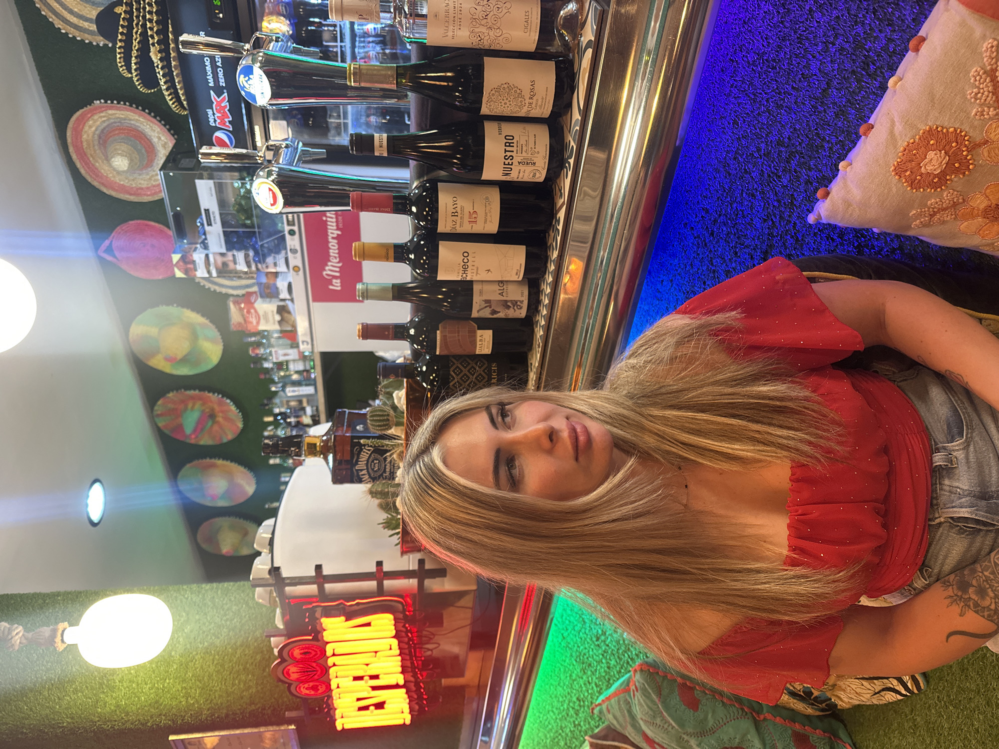 Here is a detailed description of the image:
The image depicts a woman with long, blonde hair sitting at a bar. She is wearing a red, off-the-shoulder top and denim shorts. A tattoo is visible on her left arm. Behind her, the bar is stocked with various bottles of wine and other alcoholic beverages, including Jack Daniel's.
The bar area features decorative elements such as colorful sombrero-shaped wall hangings, neon signs, and a green turf-like material on the walls and ceiling. A pillow with an embroidered design is placed on a seating area to her right. There is a sign that says "La Menorquin," indicating this is the name of the bar.
Based on the decor and the presence of the "La Menorquin" sign, it is highly likely that the location is within the city of Alicante/Alacant, Spain, as the "La Menorquin" brand has multiple locations in the area.