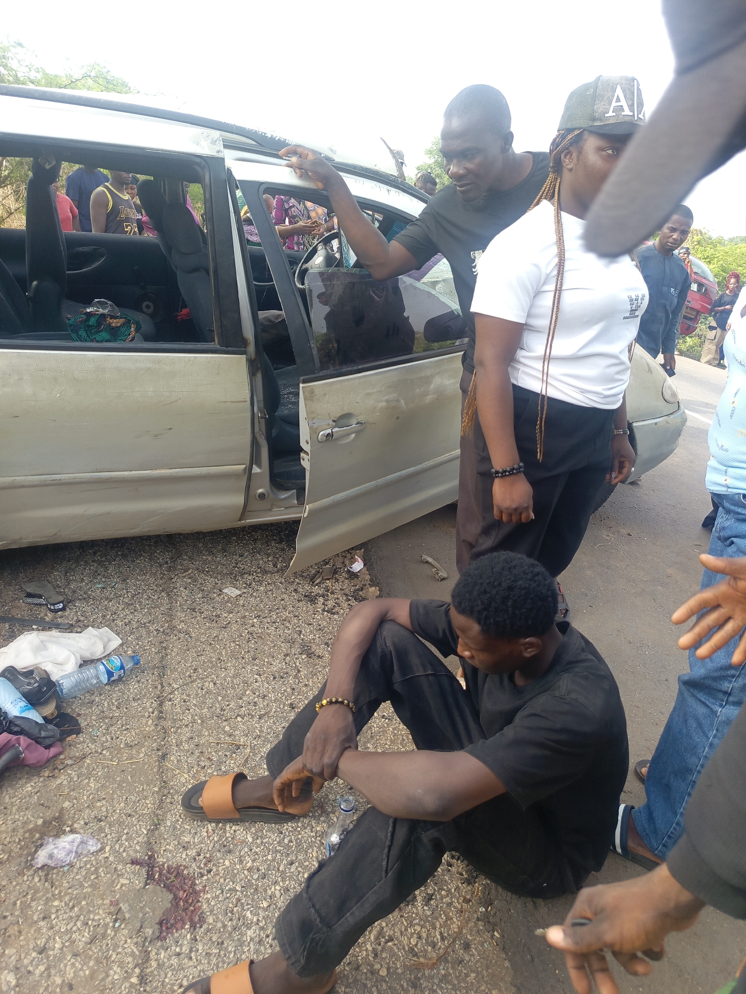 The image captures a scene on a roadside during daytime, likely following an incident involving a light-colored sedan or minivan. In the foreground, a Black man sits on the ground, dressed in a black t-shirt and black pants, wearing brown sandals. He looks down, his posture suggesting distress or injury. Near him, a reddish-brown stain, possibly blood, is visible on the gravelly surface, alongside plastic water bottles and discarded items.
The car, appearing silver or champagne, shows visible damage, including scratches and dents along its side. The front driver's side door is wide open, and the rear door is ajar, revealing the interior. The driver's side window appears to be missing. Inside the car, a person in a yellow tank top with visible text resembling "Lakers 24" can be seen.
Several other people are gathered around the vehicle. A Black man in a black t-shirt stands near the car, looking towards its interior, with his hand on the roofline. Beside him, a Black woman with long braids wears a white t-shirt, black pants, and a camouflage-patterned baseball cap featuring a stylized letter "A." Small, partially obscured text is visible on her white t-shirt. Another individual's arm is prominent in the immediate foreground, obscuring part of the scene.
In the background, more people are visible, and the setting appears to be an open area with some sparse vegetation under a bright sky. The overall atmosphere suggests concern and the aftermath of a roadside event.