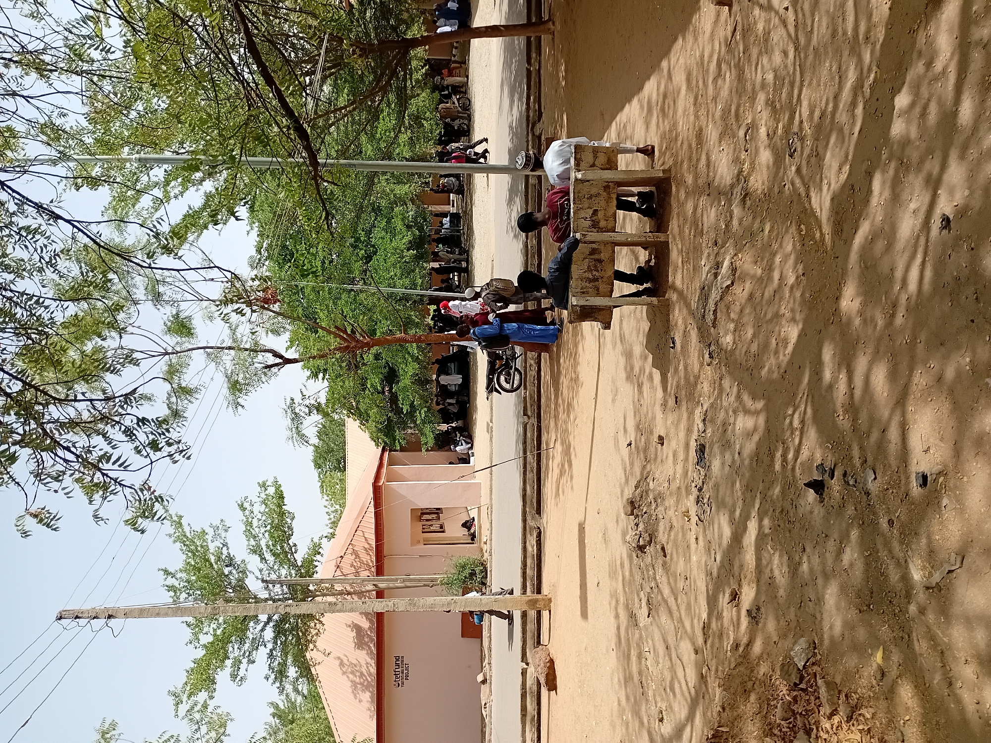 The image shows an outdoor scene in Dabawa, Nigeria. Three people are sitting on a concrete bench under the shade of a tree. A larger group of people is visible in the background, lining a street adjacent to a building with a light pink facade and a red roof. A motorcycle is parked near the group. Overhead power lines are visible between the building and the trees. The ground is dry and sandy.