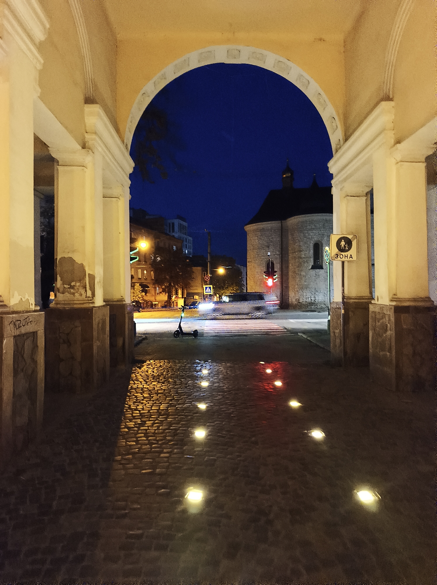The image depicts an arched passageway in Ternopil, Ukraine, at dusk. The cobblestone pavement is wet and reflects the lights embedded in the ground and from street lamps. In the distance, a traffic light shows red, and a crosswalk is visible. A pedestrian crossing sign with the Cyrillic word "ЗОНА" (ZONE) is mounted on a pole. An electric scooter is parked on the side of the road. The architecture suggests a historic European city setting. The sky is a deep blue, indicating nighttime is approaching.