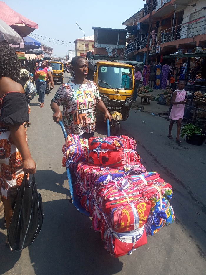 The image depicts a bustling market street scene in Nkwelle Ezunaka, Nigeria, under clear, bright daylight.
In the foreground, a woman wearing a multi-colored t-shirt with "FASHION" text is actively pushing a blue, two-wheeled handcart. The handcart is heavily laden with numerous rectangular bundles of colorful fabric, predominantly red, blue, and yellow, secured with white ties. Her gaze is directed slightly to her right. To her immediate left, partially visible, another woman wearing a black off-shoulder top and a light-colored garment with a repeating lion's head pattern holds a black plastic bag in her right hand.
In the mid-ground, a yellow three-wheeled auto-rickshaw (Keke Napep) is positioned behind the central woman and handcart. Its license plate, "PMC 424603," is partially legible. Further down the street, another yellow auto-rickshaw is visible, along with other pedestrians, including a woman in a vibrant tie-dye t-shirt. On the right side of the street, a young girl in a light pink dress stands near market stalls displaying produce such as pineapples and various items.
The background features multi-story buildings lining both sides of the paved street, some with balconies, indicating commercial or mixed-use structures. Market stalls with various goods, including fabrics and fresh produce, are visible along the sidewalks. A street light pole is present in the mid-ground on the left. Shadows are distinct on the asphalt road, consistent with strong sunlight. The overall scene is characterized by active pedestrian and commercial traffic.