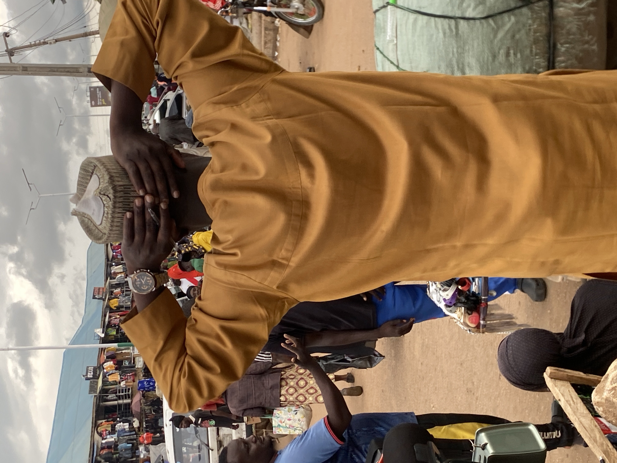 In Bukuru, Nigeria, a man is shown from behind, wearing a mustard-colored robe and a patterned cap, with his hands behind his head. He stands in what appears to be a bustling marketplace with stalls and goods displayed. In the background, other people are visible, some walking and others engaged in activities, possibly shopping or working. Vehicles, including a white car and a motorcycle, are also present in the scene. The environment seems to be an outdoor market, with a cloudy sky overhead. The overall atmosphere suggests a lively commercial area.