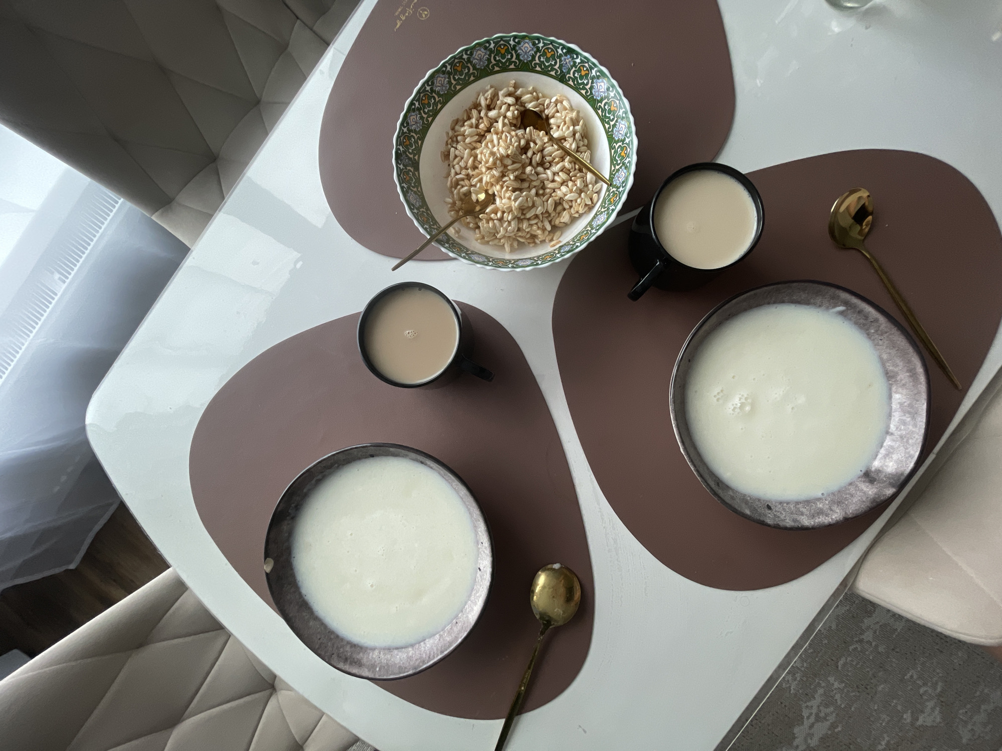 The image depicts a breakfast setting on a white table with brown placemats. Two bowls of white liquid, likely porridge or milk, are visible. One bowl is positioned on the upper left, and another on the lower left. Adjacent to the upper left bowl is a black mug filled with a light brown liquid, possibly coffee or tea, and a brass spoon. To the right of this mug is a decorative bowl filled with puffed rice cereal, also with a brass spoon. A second black mug, containing a similar light brown liquid, is situated on the lower right, below the puffed rice cereal bowl. The scene appears to be indoors, with glimpses of upholstered chairs and a window with blinds in the background. There are no people visible in the image. The location is stated as Podshivalovo, Russia.