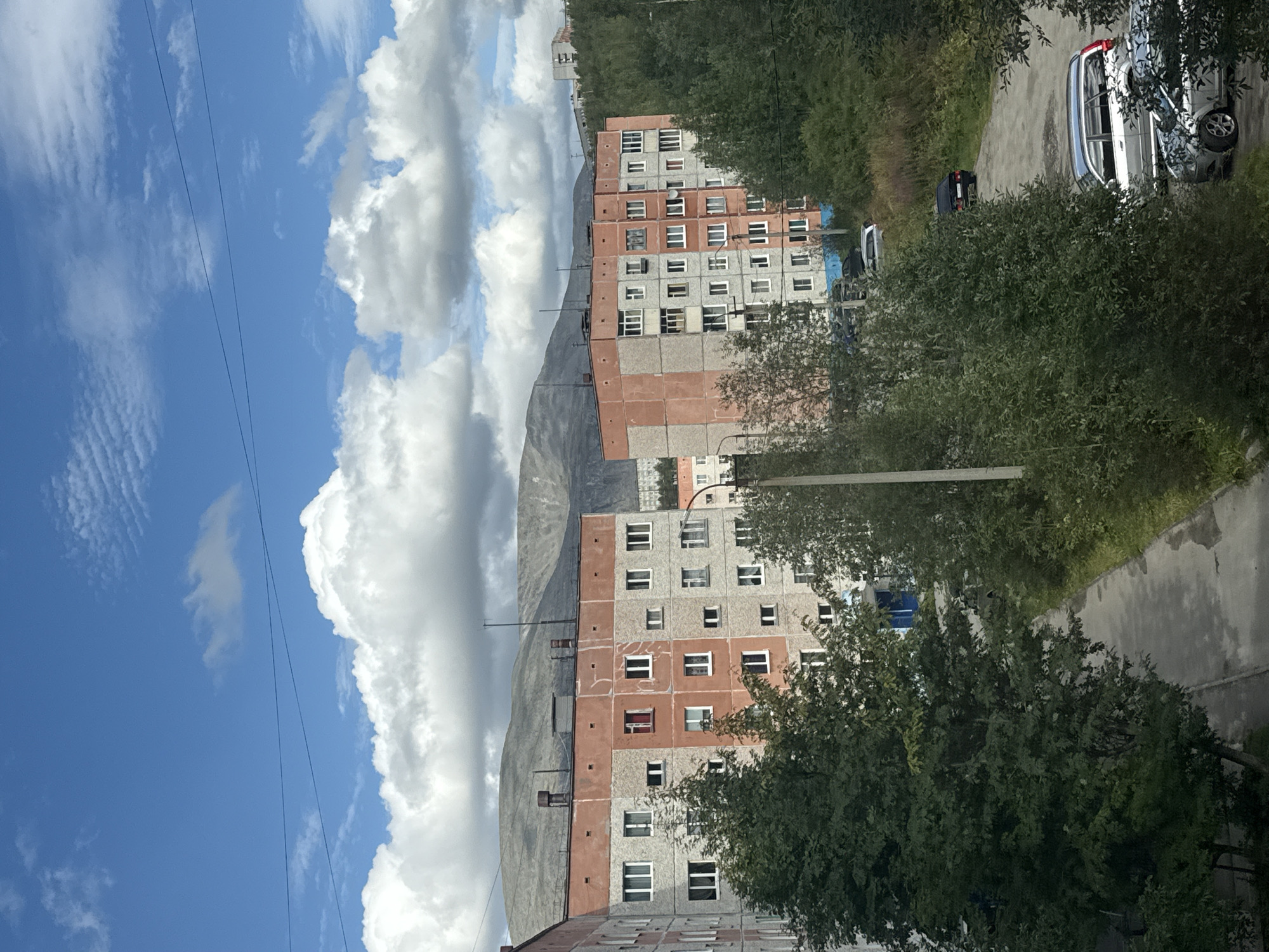The image captures a view of apartment buildings in Kirovsk, Russia, under a partly cloudy sky with distant mountains. The apartment buildings are constructed with concrete panels, with some sections painted in a peachy-orange color. The windows are predominantly white, with some balconies visible.
In the foreground, there are trees and vegetation, suggesting a residential area with some greenery. A paved path or road runs through the scene, with several parked cars visible, including a silver station wagon and other dark-colored vehicles. The architecture and the presence of mountains in the background are characteristic of Kirovsk, a city known for its mining industry and proximity to the Khibiny Mountains. The overall impression is a somewhat stark but functional urban landscape in a northern environment.