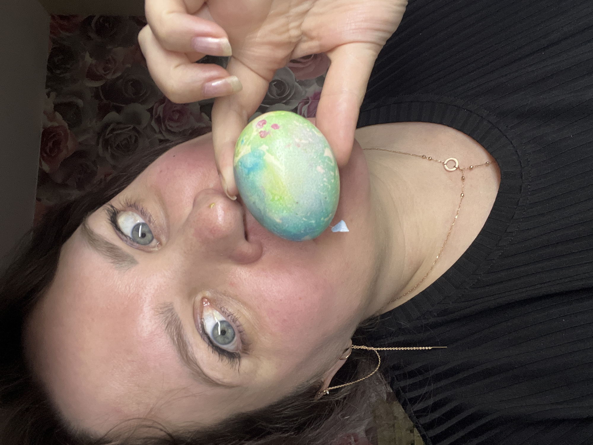 A woman with light skin, blue eyes, and dark hair is holding a painted Easter egg near her face. The egg is light green and blue with pink spots. She is wearing a black shirt and a gold necklace. The background includes a wall with a floral pattern. The location is unknown, and there is no indication of Noviy, Russia.