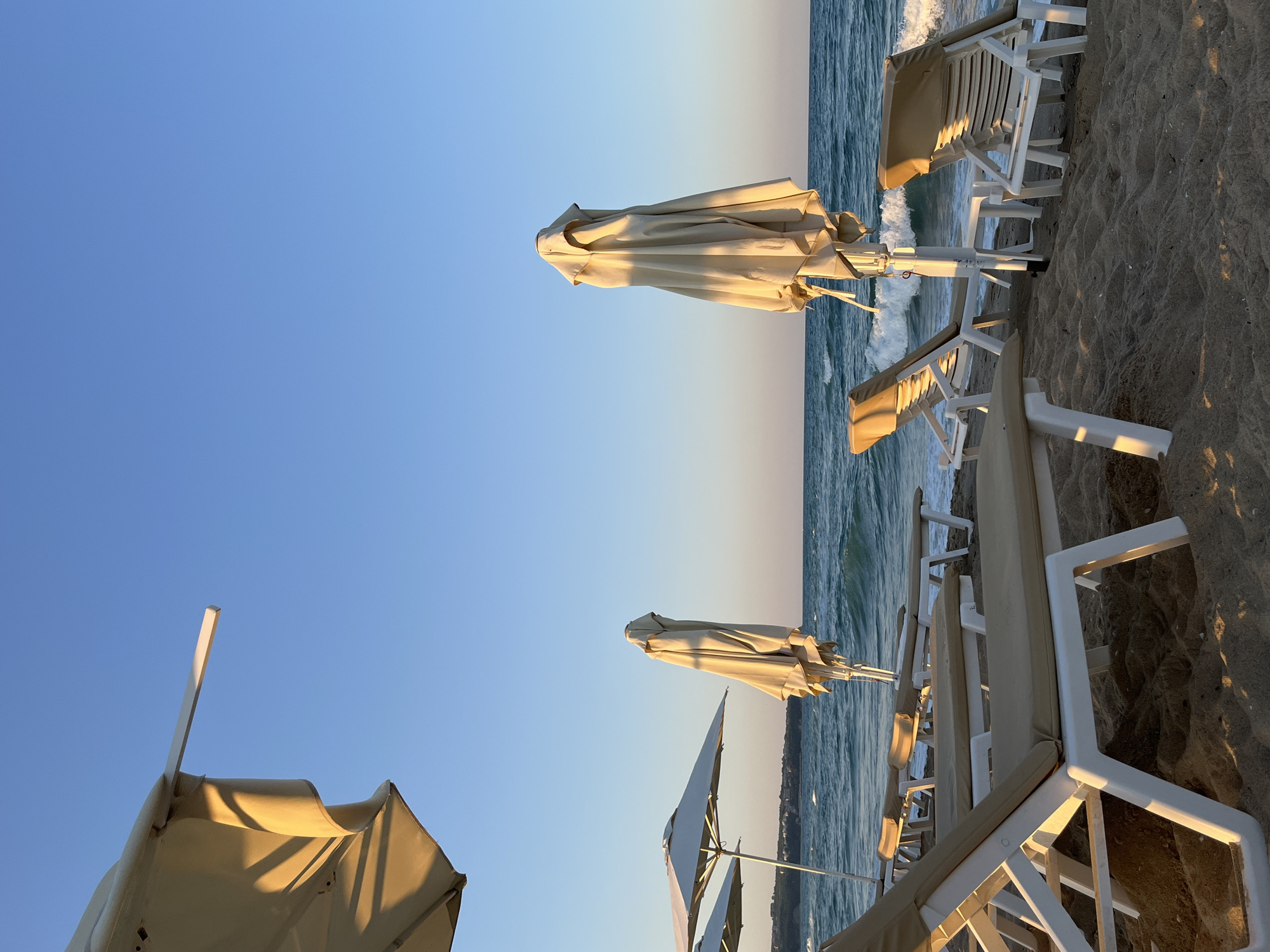 The image depicts a serene beach scene in Obzor, Bulgaria, during the golden hour. Several white plastic sun loungers with beige cushions are arranged on the sand. Some sun loungers are empty, while others are occupied by unseen individuals or are positioned in a way that suggests recent use.
In the background, the calm sea with gentle waves stretches towards the horizon. The sky is a clear, pale blue, transitioning to a warmer hue near the horizon, indicating either sunrise or sunset. Several closed beige umbrellas are placed between the sun loungers, providing shade.
The overall impression is one of tranquility and relaxation, typical of a beach resort. The lighting suggests a peaceful atmosphere, with soft shadows cast by the loungers and umbrellas on the sand. The scene appears to be devoid of any immediate activity or human interaction beyond the static arrangement of beach furniture.
