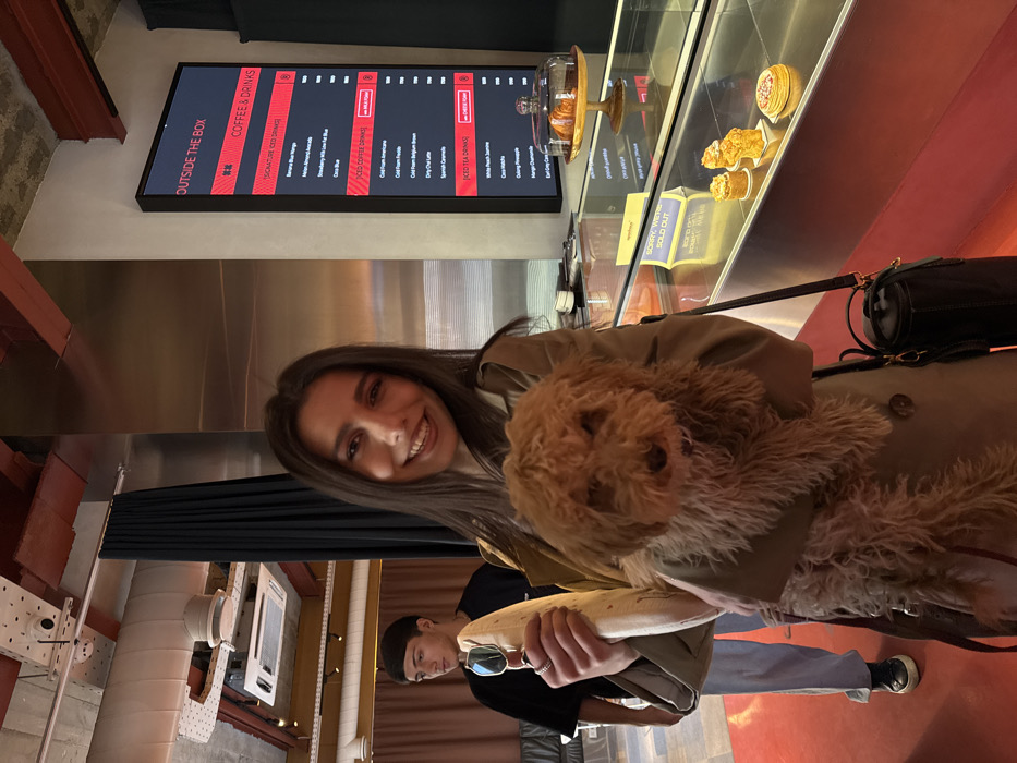 A young woman, smiling and looking directly at the camera, stands in the foreground inside the "OUTSIDE THE BOX" cafe in Yerevan, Armenia. She holds a small, light brown, curly-haired dog in her left arm. In her right hand, she holds a light-colored, elongated object, possibly a wrapped baguette, and a pair of sunglasses. She is wearing a light brown trench coat and a black cylindrical handbag with a shoulder strap.
In the background, to the left of the woman, a young man with dark hair, wearing a dark short-sleeved shirt and light-colored pants, is partially visible. To the woman's right, a vertical black digital display board lists "OUTSIDE THE BOX COFFEE & DRINKS" with various beverages and prices. Below the menu board, a glass display case holds baked goods, including a croissant under a glass dome on a wooden stand and several individual muffins or cakes. A purple and white sign inside the display case reads "SORRY, WE'RE SOLD OUT."
The cafe's interior features an industrial aesthetic with an exposed concrete ceiling, visible metal ducts, and recessed lighting. A black curtain hangs vertically in the mid-ground, and a red floor is prominent throughout the lower portion of the scene.