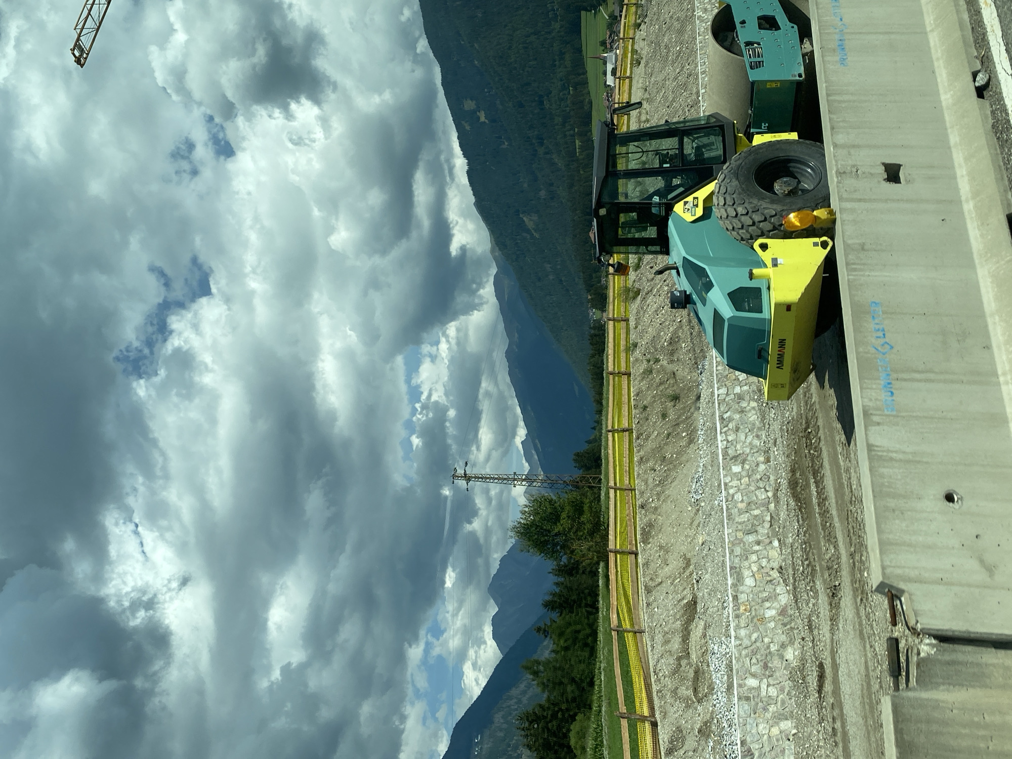 An Ammann asphalt compactor, painted teal and yellow, is positioned on a dirt construction site in Rasen-Antholz - Rasun-Anterselva, Italy. The compactor has a large, textured tire in the front and a smooth roller in the back. The site is characterized by exposed earth, gravel, and a low stone wall. In the background, a yellow safety fence borders a slightly elevated area. Beyond the fence, a field with some crops is visible, followed by a dense line of evergreen trees. Further in the distance, majestic mountains rise under a dramatic sky filled with dark, textured clouds. A tall electrical pylon with power lines is situated between the trees and the mountains. A construction crane's boom is visible in the upper right corner against the cloudy sky. In the far distance, a white church with a red roof stands on a green hillside. A concrete barrier, partially obscuring the compactor, is in the foreground, with blue spray-painted markings on its surface. The overall scene depicts an ongoing construction project in a mountainous, rural landscape.