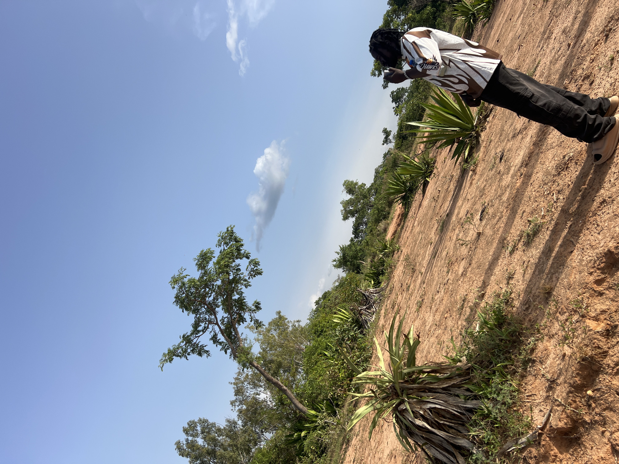 A person with dark hair in dreadlocks, wearing a white and brown patterned long-sleeved shirt and black pants, stands on a reddish-brown, uneven dirt surface in what appears to be a rural area. The person is looking down, seemingly focused on something in their hands, possibly a phone. Scattered vegetation is present, including various plants and trees. In the distance, a blue sky is visible with some clouds. The location is Fobur, Nigeria.