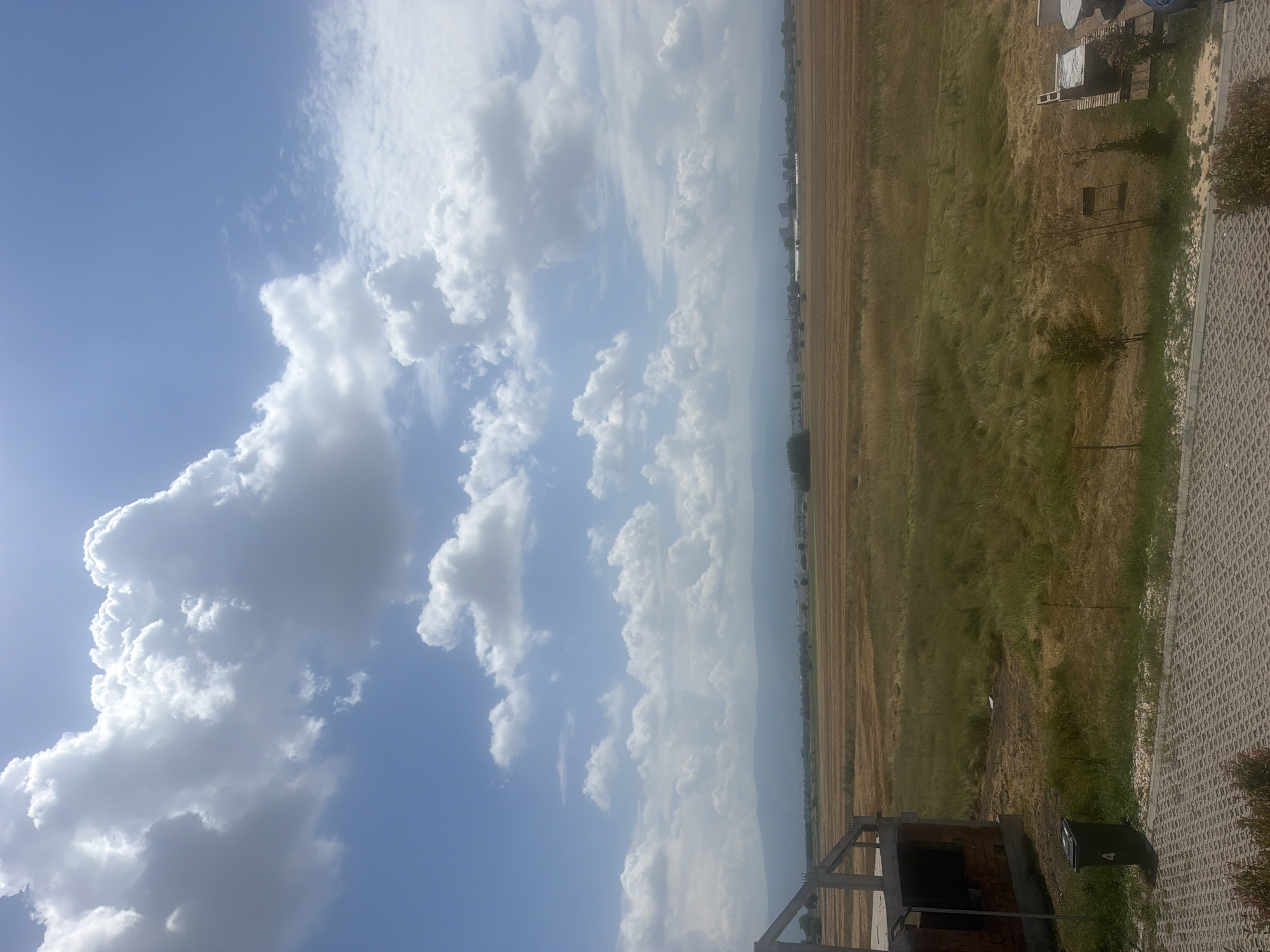 The image presents a wide outdoor landscape in Skutare, Bulgaria, under a clear blue sky populated by numerous large, white cumulus clouds, indicating a bright, sunny daytime environment. In the immediate foreground, a grey interlocking paver patio extends across the bottom, bordered by a strip of dry grass and a few small, sparse shrubs. To the left, a partially constructed building with exposed concrete framing and red brick walls is visible, adjacent to a black waste bin marked with the number "4." To the right, a segment of an outdoor seating area featuring a covered table and chairs is partially visible. The midground consists of extensive, flat agricultural fields displaying a mixed texture of golden-brown stubble from harvested crops and patches of taller, lighter green grasses. The distant background reveals a long, hazy mountain range beneath a band of faint urban structures. No people, animals, or active interactions are present in the scene.