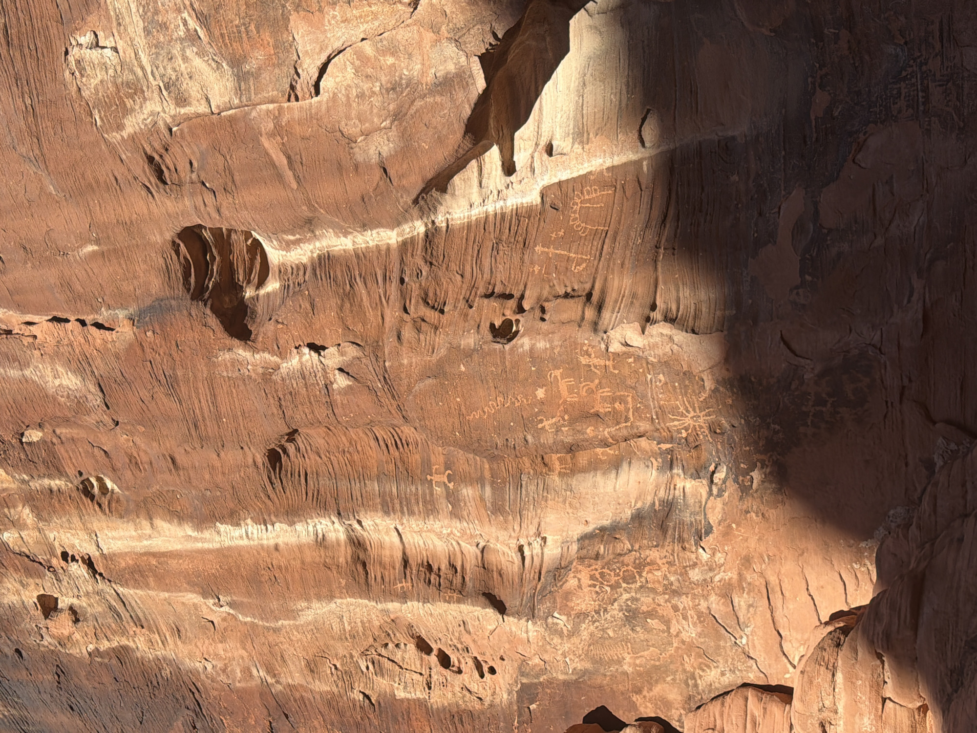 The image shows a rock wall, likely sandstone, with various petroglyphs carved into the surface. The rock is reddish-brown, and there are areas of light and shadow. The petroglyphs are faint and appear as simple line drawings. The environment suggests an outdoor setting, possibly in a desert or canyon area. Based on the location provided, this is likely to be a petroglyph site in the Moapa Valley, United States.
