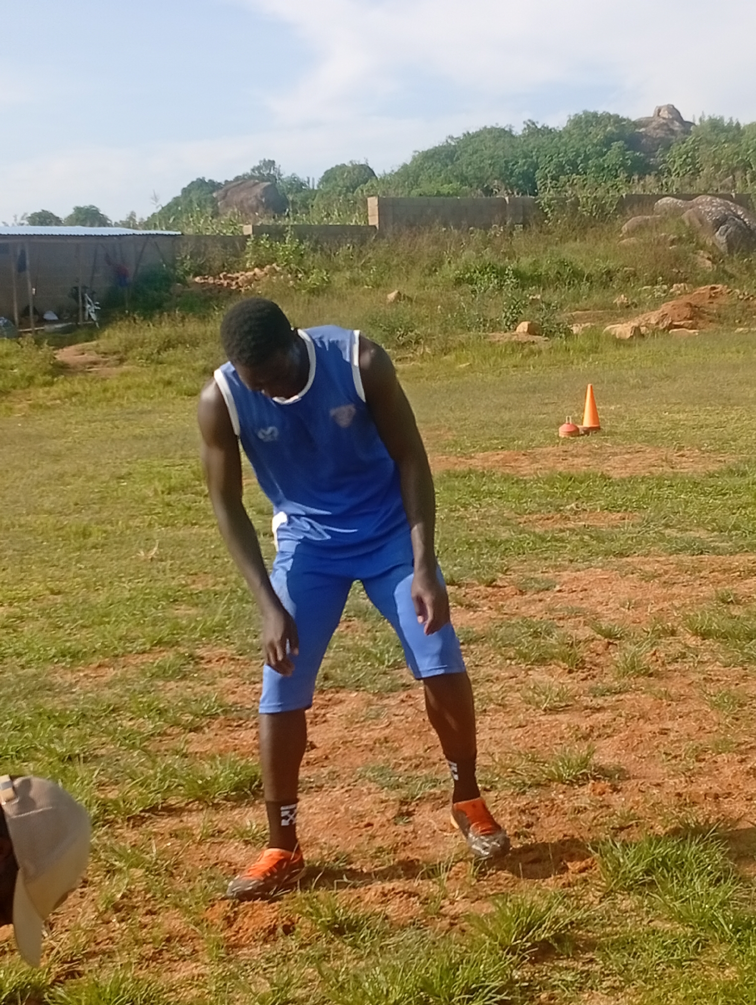 In a dusty, grassy field, a young Black man wearing a blue athletic uniform, including a tank top and shorts, is engaged in what appears to be a warm-up or stretching exercise. His head is bowed, and his arms are bent at the elbows, with his hands resting on his thighs. He is wearing orange athletic shoes with black and silver accents, and black socks with a white logo. The background features a slightly overgrown landscape with scattered rocks, a rough concrete wall, and a corrugated metal structure. In the middle ground, an orange traffic cone is visible on the grass. The sky is clear and blue, suggesting fair weather. The scene is likely located outdoors in a less developed area, possibly on the outskirts of the city of Jos, Nigeria, given the arid terrain and simple structures.