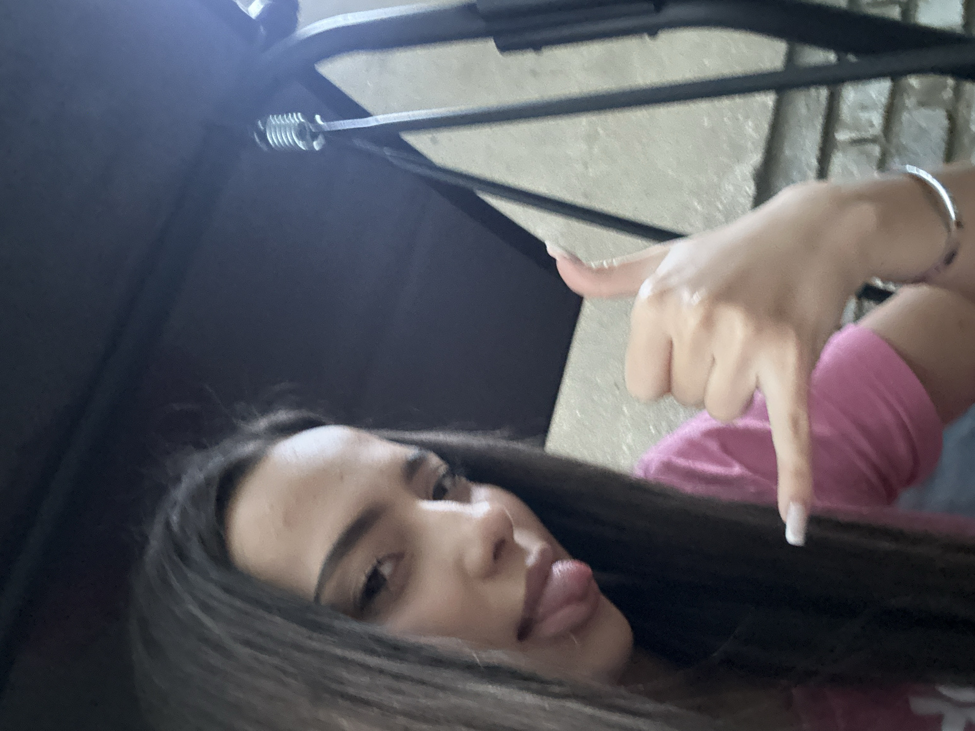 A close-up, slightly angled shot depicts a person with dark hair sticking out their tongue. The person is wearing a pink shirt. The person is making a hand gesture resembling a thumbs-down, with the finger pointing downward. A metal object and a spring are visible in the upper portion of the frame. The background appears to be a stone or concrete surface. The location and city are unknown.