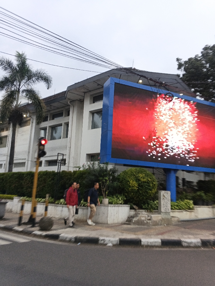 This image captures a daytime urban scene in Bandung City, Indonesia, beneath an overcast sky. In the foreground, an asphalt road with a visible crosswalk and a black and white striped curb transitions to a concrete sidewalk on the right. A yellow-poled traffic light with an illuminated red signal is positioned on the left. Three adult males are walking from left to right on the sidewalk; one is wearing a red long-sleeved shirt, another is partially obscured in a dark shirt, and the third is wearing a dark blue polo shirt and light-colored pants, carrying a dark backpack. A spherical grey concrete object rests on the sidewalk near the traffic light. A low concrete planter, containing green foliage and a white rectangular stone marker, separates the sidewalk from a large, blue-framed LED digital billboard on the right. The billboard displays a vibrant red background with numerous scattered white light patterns, resembling fireworks. Behind the billboard and to its left, a multi-story white building with a dark roof and multiple windows is visible. A tall palm tree stands on the far left, and a dense green tree canopy is visible on the far right. Multiple black utility lines crisscross the upper portion of the frame against the sky.