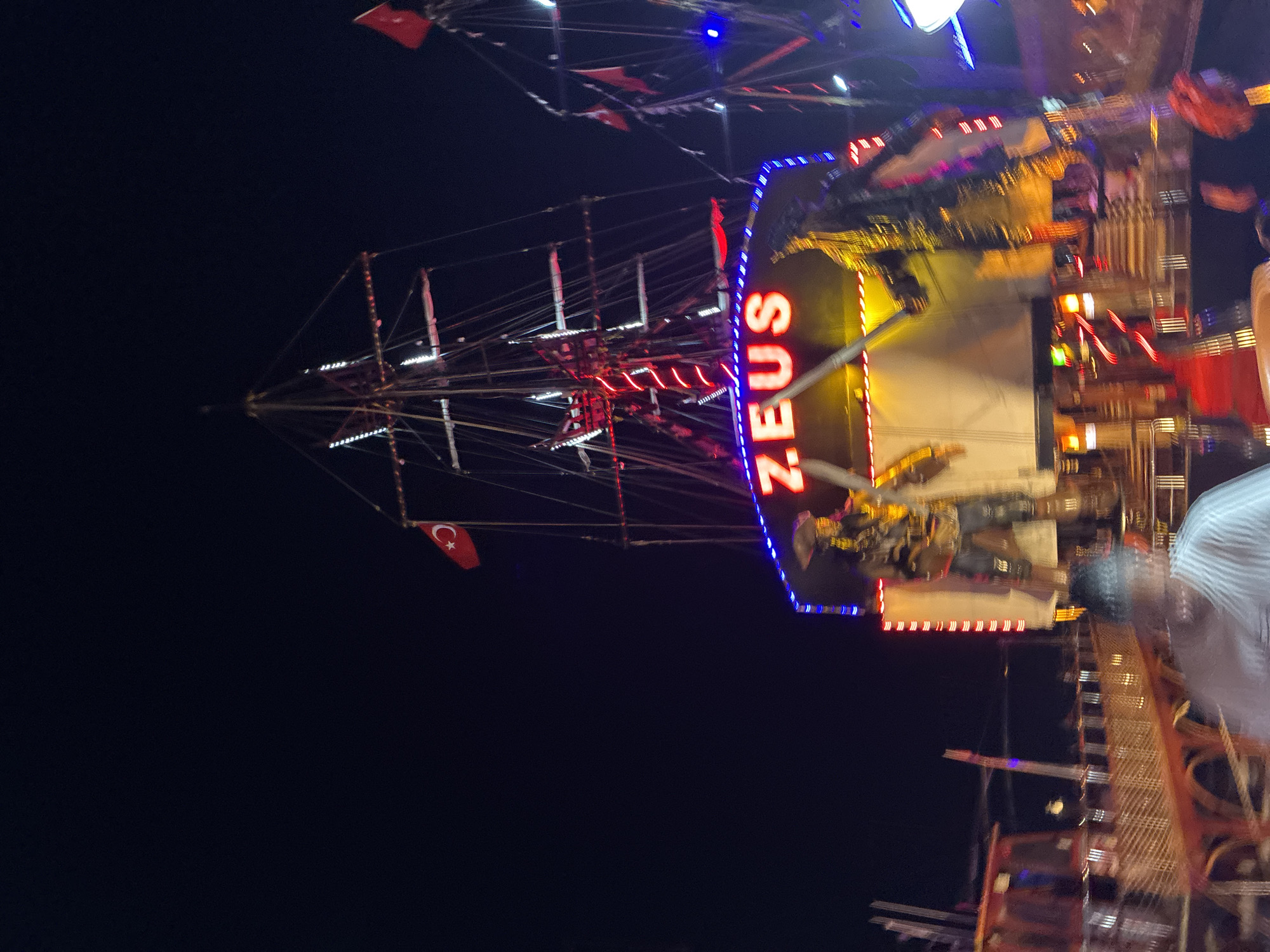 A blurry, night-time, low-angle, medium shot captures a decorated, stationary pirate ship at what appears to be an amusement park or waterfront attraction in Alanya, Turkey. The ship is adorned with bright, colorful LED lights in blue, red, and yellow, outlining its structure and the prominent word "ZEUS" in large, red neon letters. Two large pirate figures, one with a sword, stand on a platform at the front of the ship. The ship's masts and rigging are visible against the dark night sky, with several red flags flying, including a Turkish flag. In the foreground, a person in a striped shirt is blurred, as are other indistinct figures and structures of the amusement area, suggesting movement or a quick capture of the scene.