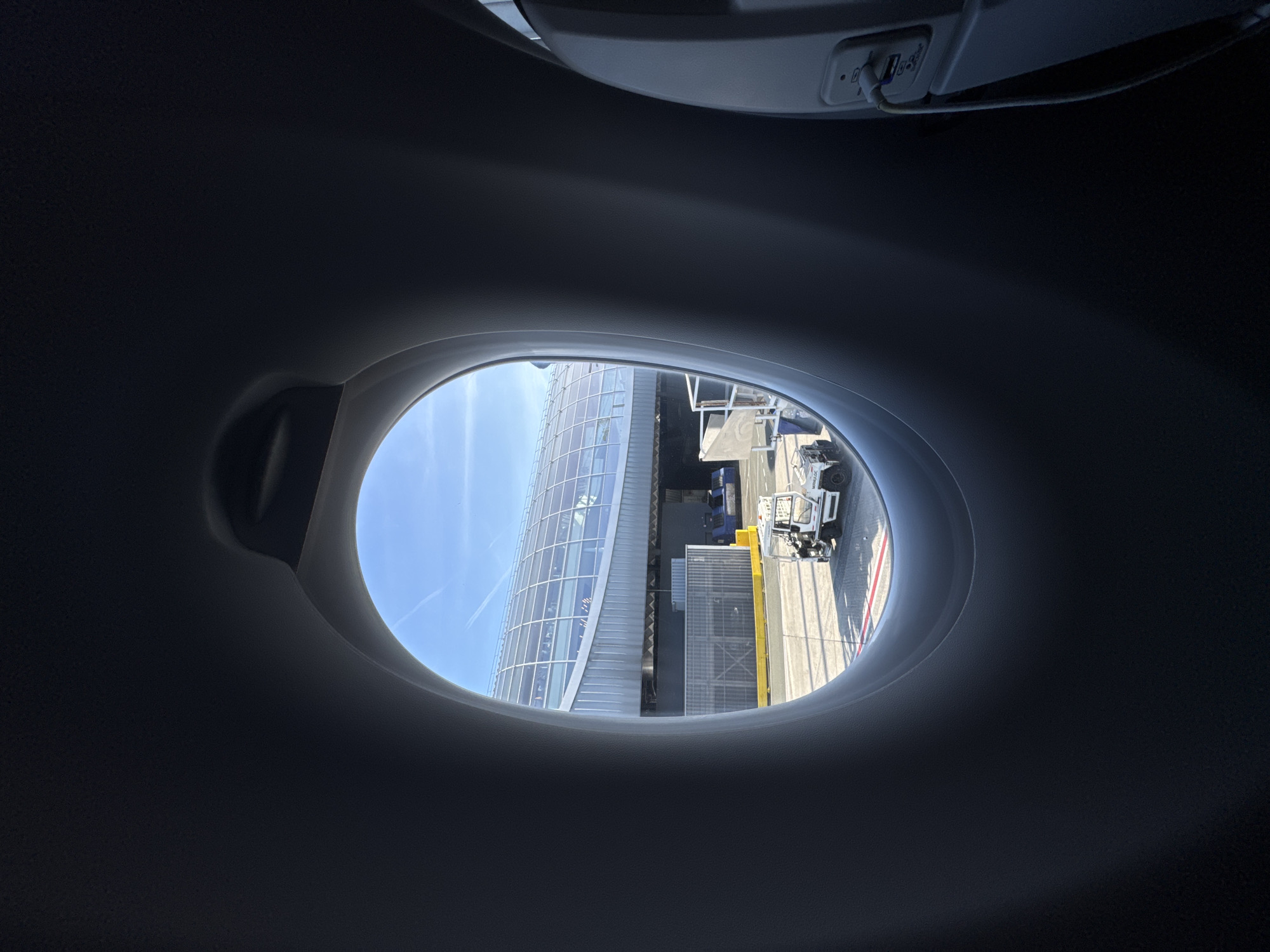 View from an airplane window looking out at the airport terminal at Charles de Gaulle Airport in Le Mesnil-Amelot, France. A modern glass-walled terminal building is visible, along with airport vehicles and equipment on the tarmac. The sky is blue. The view is framed by the airplane cabin interior, including a USB charging port.