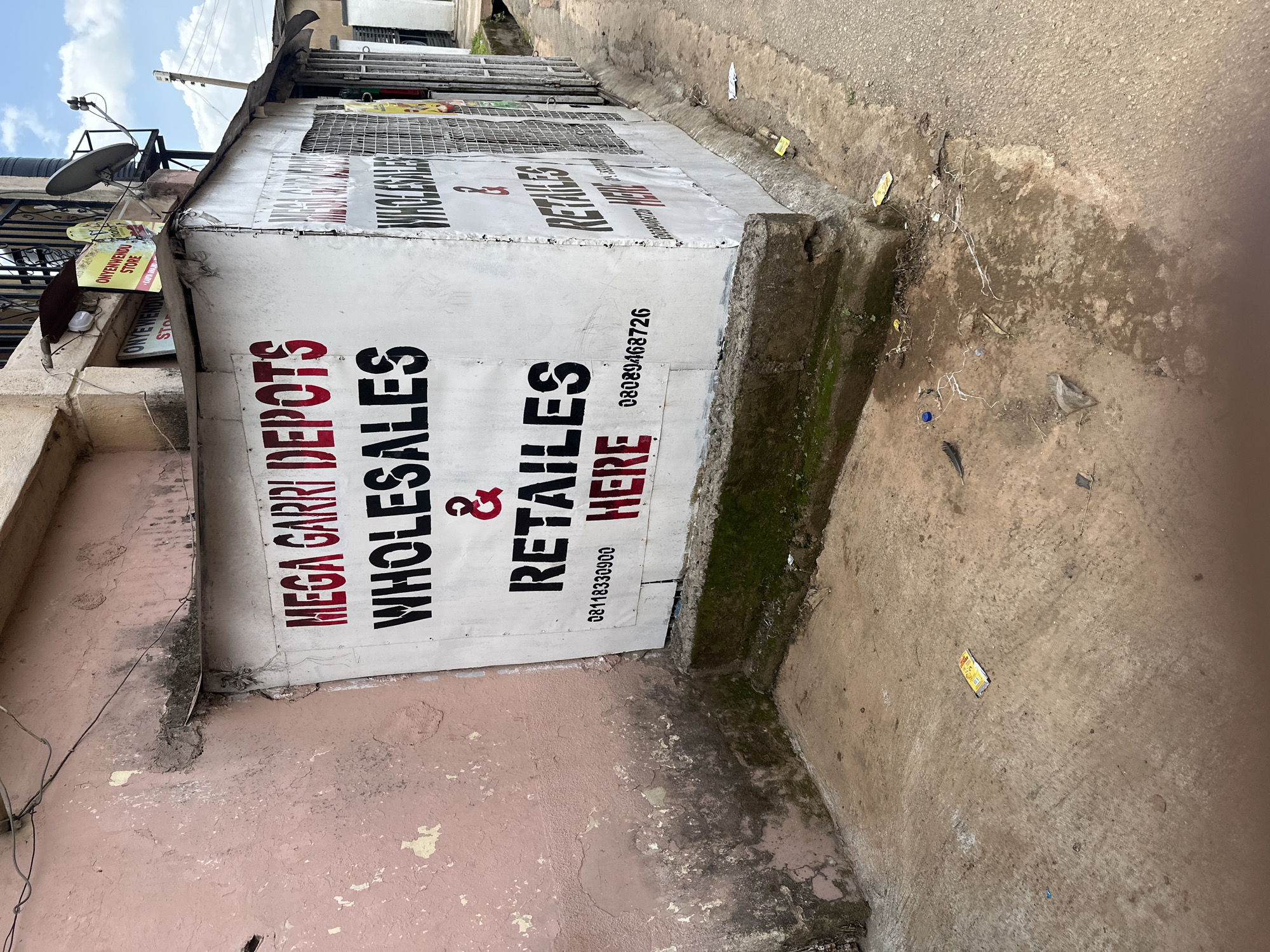 The image shows a dilapidated building with a sign that reads "MEGA GARRI DEPOTS WHOLESALES & RETAIL ES HERE". The sign is painted in red and black letters on a white background. The building appears to be made of corrugated metal and is situated on a street with a concrete sidewalk. There are some signs of wear and tear on the building, including peeling paint and moss growth on the concrete base. The street is paved with asphalt and there is some debris on the ground, including a yellow packet and a blue bottle cap. The sky is cloudy and the overall lighting suggests it is daytime. The scene depicts an outdoor commercial establishment in what appears to be a less developed urban area.