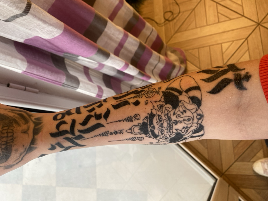 An overhead close-up image shows the tattooed left forearm of an individual in Kunje, Montenegro. The arm features several black ink tattoos: a faded skull on the upper arm, multiple lines of calligraphic script, and a prominent central design depicting a crowned, horned figure with wide eyes and bared teeth, surrounded by smaller symbolic lines. Towards the wrist, a stylized black tattoo resembling flame-like shapes is partially visible, with a red knitted cuff obscuring the lower forearm. The background consists of a light-colored window or door frame and a light wooden floor with a geometric pattern on the left, and a vertically striped curtain with white, purple, and gray bands on the right.
