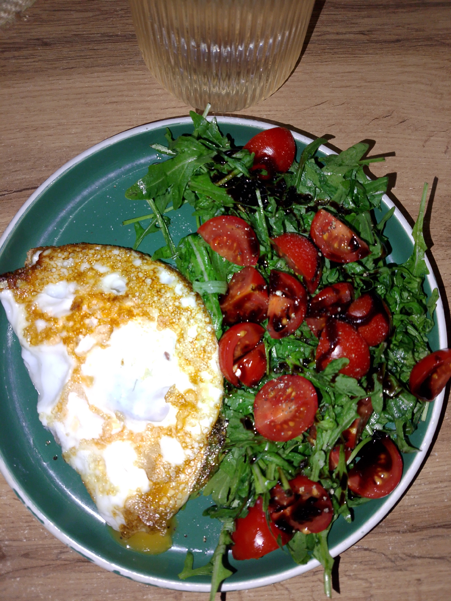 A top-down shot shows a green plate with a fried egg and a salad of arugula and halved cherry tomatoes, drizzled with balsamic glaze, on a wooden table. Above the plate, partially visible, is a clear glass bowl. The plate is predominantly green with a thin white border. The egg is fried with crispy edges and a runny yolk. The salad is composed of fresh green arugula and bright red, halved cherry tomatoes, glossy from the balsamic glaze. The table has a brown, wood-grain surface. The lighting is somewhat dim. There is no evidence suggesting the location of the photo is in Izhevsk, Russia.
