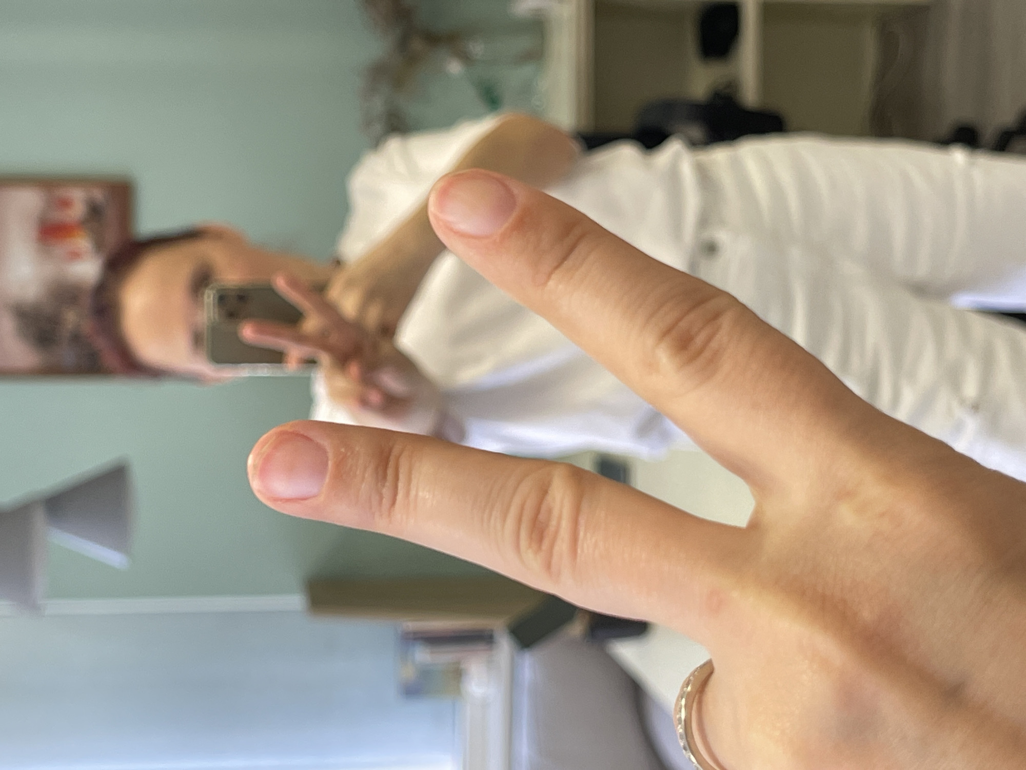 A person is reflected in a mirror, holding up a hand in a peace sign gesture. The person is wearing white clothing. A gold ring is visible on the person's hand. The environment includes a light green wall and a shelf with various items. The location in Batumi, Georgia is not discernible from the image.