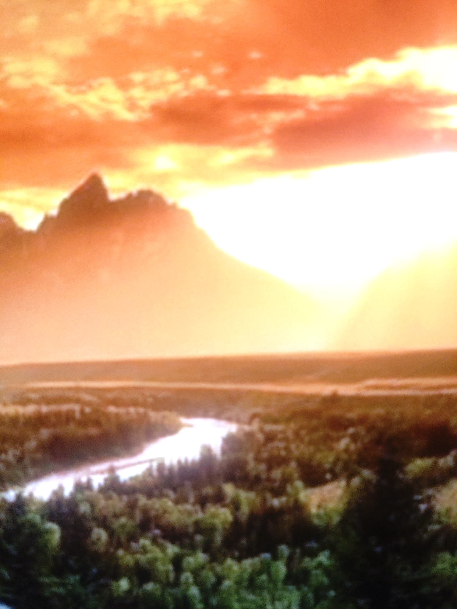 The image depicts a dramatic sunset or sunrise over a mountainous landscape. The sky is filled with vibrant orange and yellow hues, with bright white light emanating from the right side, likely the sun. Silhouetted against this light are the jagged peaks of mountains. In the foreground, a winding river flows through a valley covered with lush green trees, suggesting a natural, undeveloped area. The overall impression is one of natural beauty and vastness. There is no indication of the city of Armavir, Russia, or any urban elements in this image.