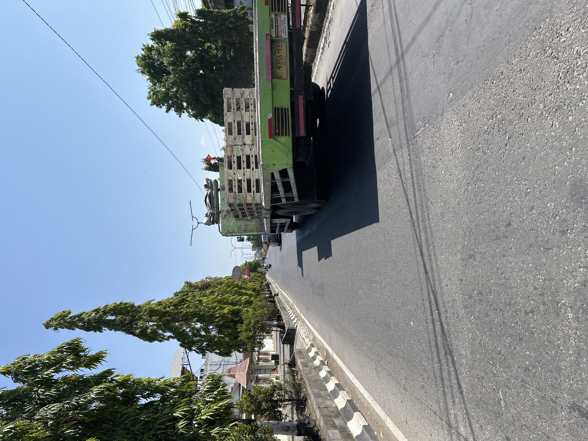 This is a ground-level, outdoor, eye-level shot of a paved road in Bulakrejo, Indonesia. A large, light green flatbed truck is moving away from the camera on the right side of the road, carrying a load of light-colored wooden pallets. The truck has a yellow license plate with the characters "F 9356 FE". On the back of the truck, there is a red and white flag. There are trees lining the left side of the road, and a building with a red-tiled roof is visible behind them. The sky is clear and blue. Other vehicles and motorcycles are also visible on the road in the distance. The road has white lane markings and some cracks in the asphalt. The scene is well-lit by sunlight, creating shadows on the road.