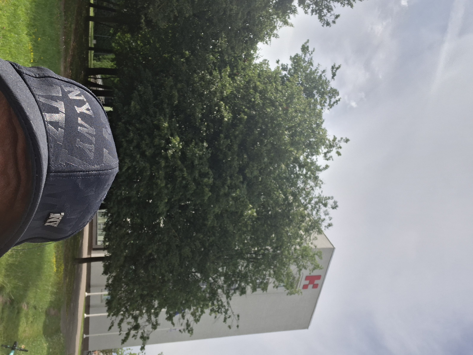 The image shows a close-up of a person wearing a navy blue baseball cap with a metallic "NY" logo. The cap is angled to show the front. In the background, there is a large, leafy green tree. Behind the tree and to the right, a modern white building is visible with a red symbol that resembles an "H" on its facade. The building appears to be a hospital or medical facility, given the common association of the red cross symbol with healthcare, although this symbol is a stylized "H". The setting is outdoors, with a grassy area and a pathway visible in the foreground and middle ground. The sky is overcast with some lighter patches, suggesting it might be a cloudy or hazy day. The image is oriented vertically, and appears to have been taken during daylight hours. The context provided indicates the location is Kaunas, Lithuania. There are no discernible people in the image other than the implied wearer of the cap. The overall impression is a casual, everyday scene.