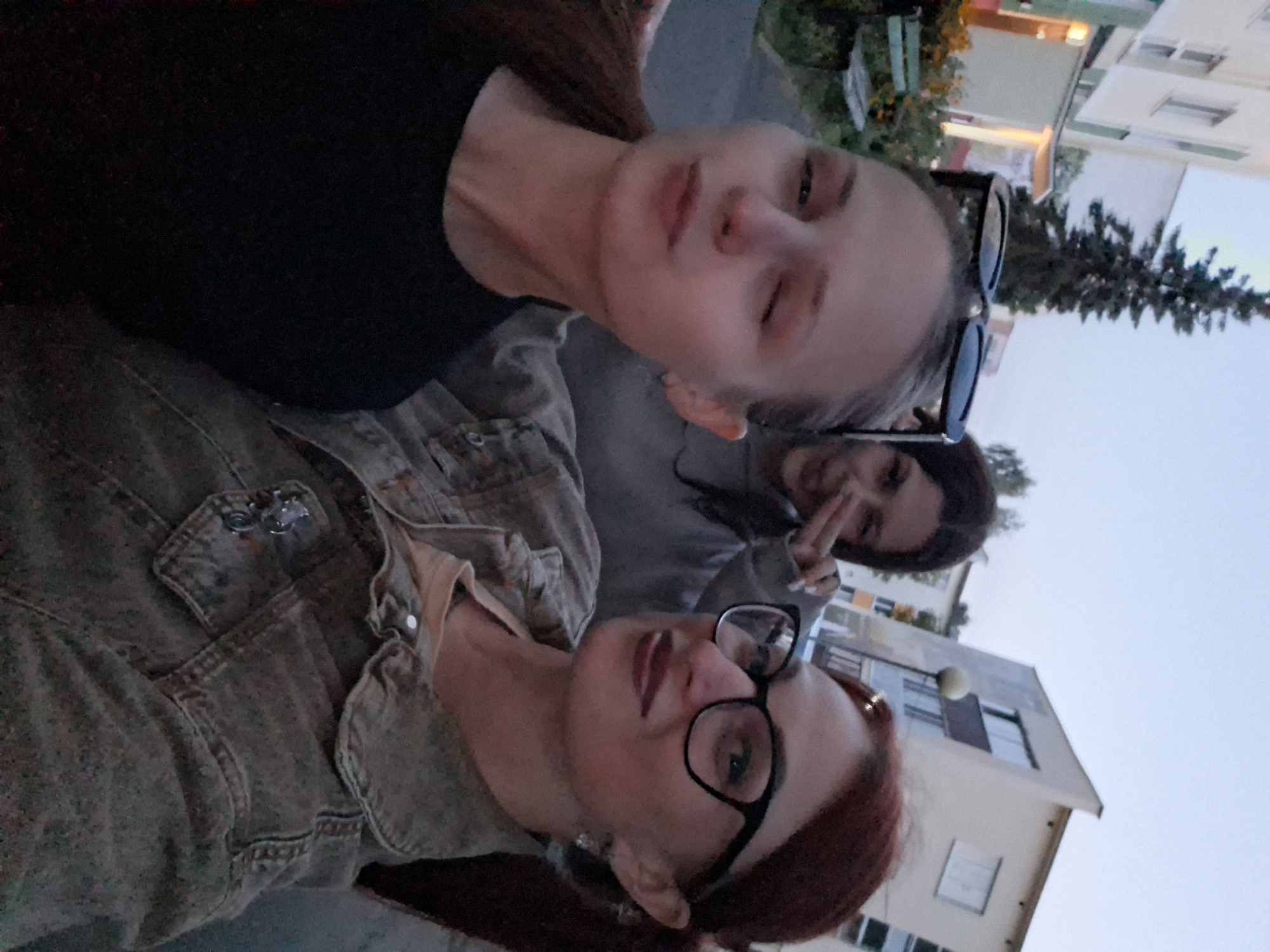 Three women are taking a selfie outdoors. The woman in the foreground on the left is looking at the camera with one eye closed and her lips slightly pursed, wearing a black t-shirt. The woman in the middle is making a peace sign with her fingers and smiling, wearing a grey hoodie. The woman on the right is wearing glasses and a denim jacket over a yellow shirt, also smiling. They are standing in what appears to be a residential area, with a building visible in the background and a tall evergreen tree to the left. The lighting suggests it is either early morning or late evening. The location is identified as Myadzyel, Belarus.