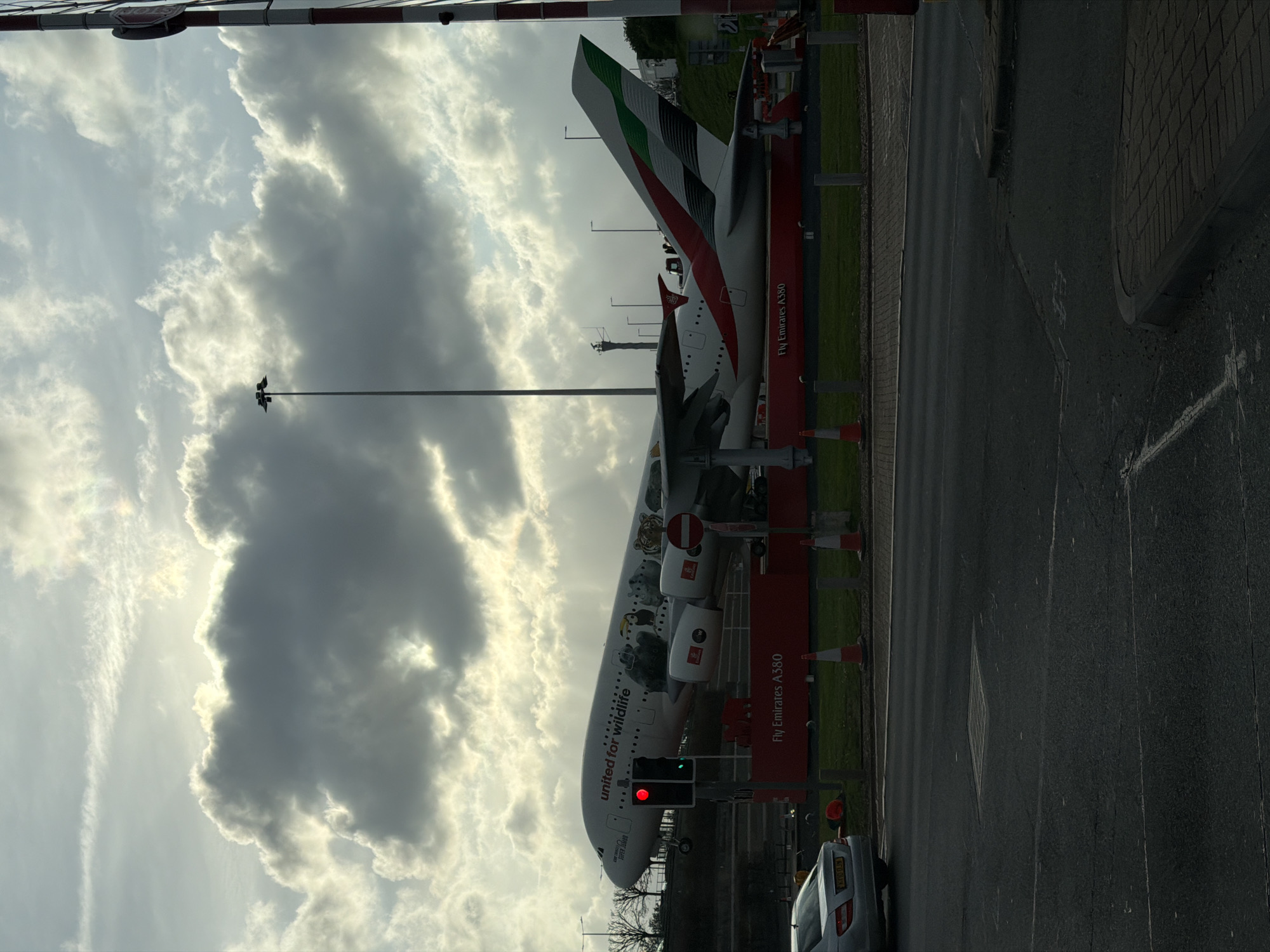 The image shows an Emirates A380 aircraft with "United for Wildlife" livery parked on a stand or a display area, possibly near a road. The tail fin is visible, with the Emirates flag design. Traffic lights are present, showing red, and a silver car is partially visible in the lower right corner. The aircraft is positioned adjacent to a green area. The sky above is cloudy with the sun partially obscured. The image was taken near London Heathrow Airport; this is evidenced by the aircraft's "Fly Emirates A380" livery and by the proximity of roads and traffic signs typical of airport perimeters in the United Kingdom. A road barrier is also seen at the top of the image.