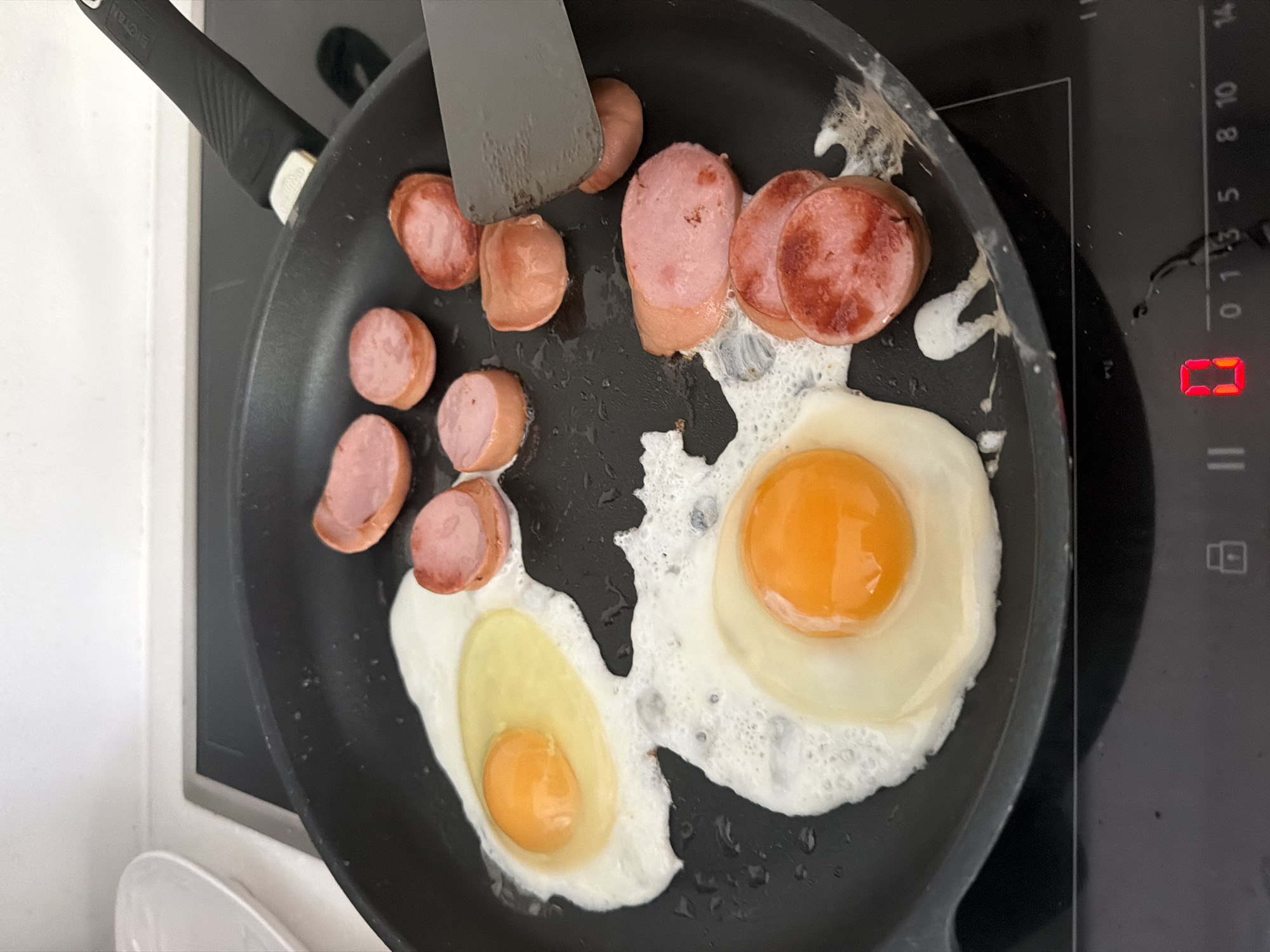 In Baloži, Latvia, a black frying pan is on a black induction stovetop. The pan contains two fried eggs, one with a fully cooked white and runny yolk, and another with a partially cooked white and runny yolk. There are several slices of cooked sausage in the pan, some appearing browned and crispy. A metal spatula is in the pan, positioned to flip a slice of sausage. The stovetop display shows a red "0" indicating the heat level. The background is out of focus, showing a white countertop and a white plate.