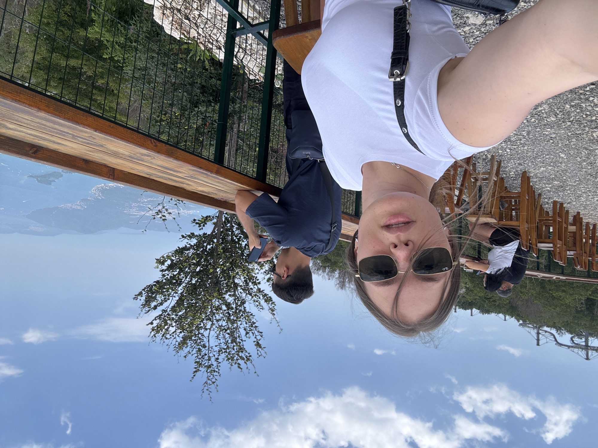 This is a selfie taken outdoors by a woman with long brown hair, wearing sunglasses and a white t-shirt. She is sitting at a wooden table. Behind her, a man wearing a dark blue t-shirt and a dark bag slung across his chest is visible, holding a phone. Further in the background, there are more people sitting at tables and chairs, with wooden fencing and greenery. The scene appears to be a scenic overlook, with a view of a valley and a distant body of water under a blue sky with scattered white clouds. A roller coaster track is visible in the far background. The overall impression is a relaxed, scenic outing on a sunny day.